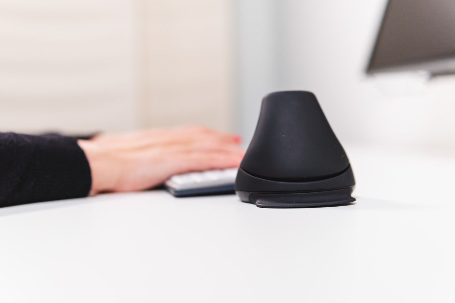Un mouse ergonomico nero è su una scrivania bianca, vicino a una tastiera e alle mani.