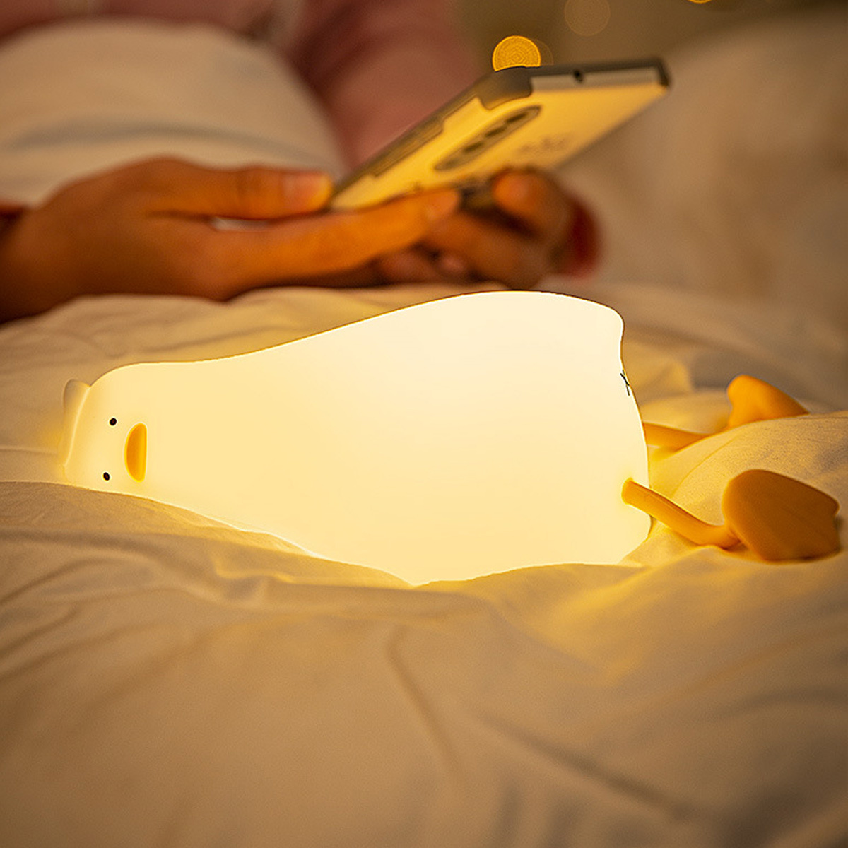 Weißes Enten-Nachtlicht auf einem Bett. Person, die im Hintergrund ein Telefon benutzt. Warmes Licht.