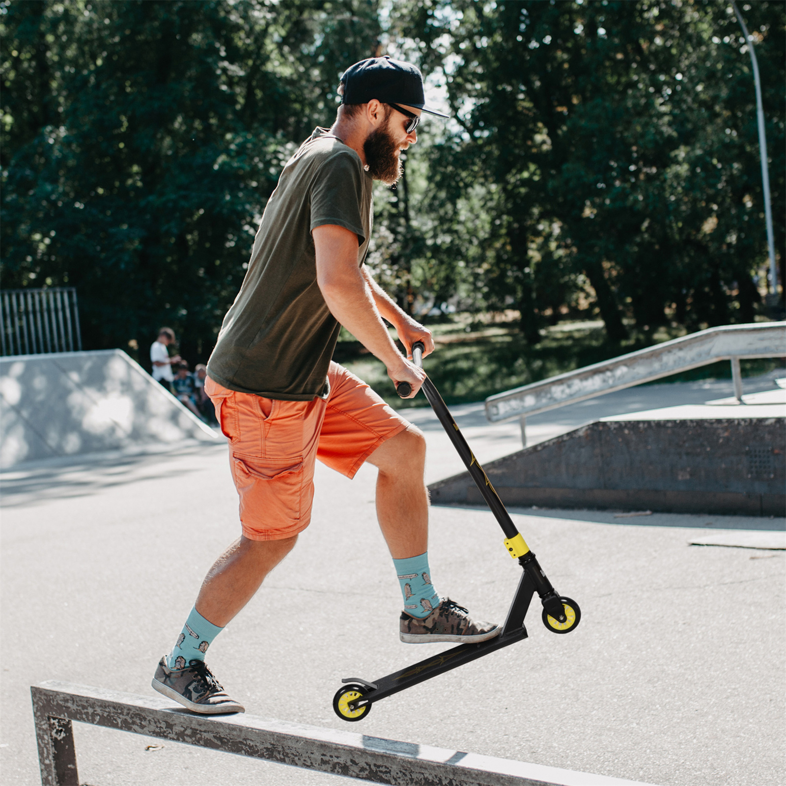 Mann auf Roller in Skatepark, kurz davor, auf Schiene zu fahren. Bäume im Hintergrund.