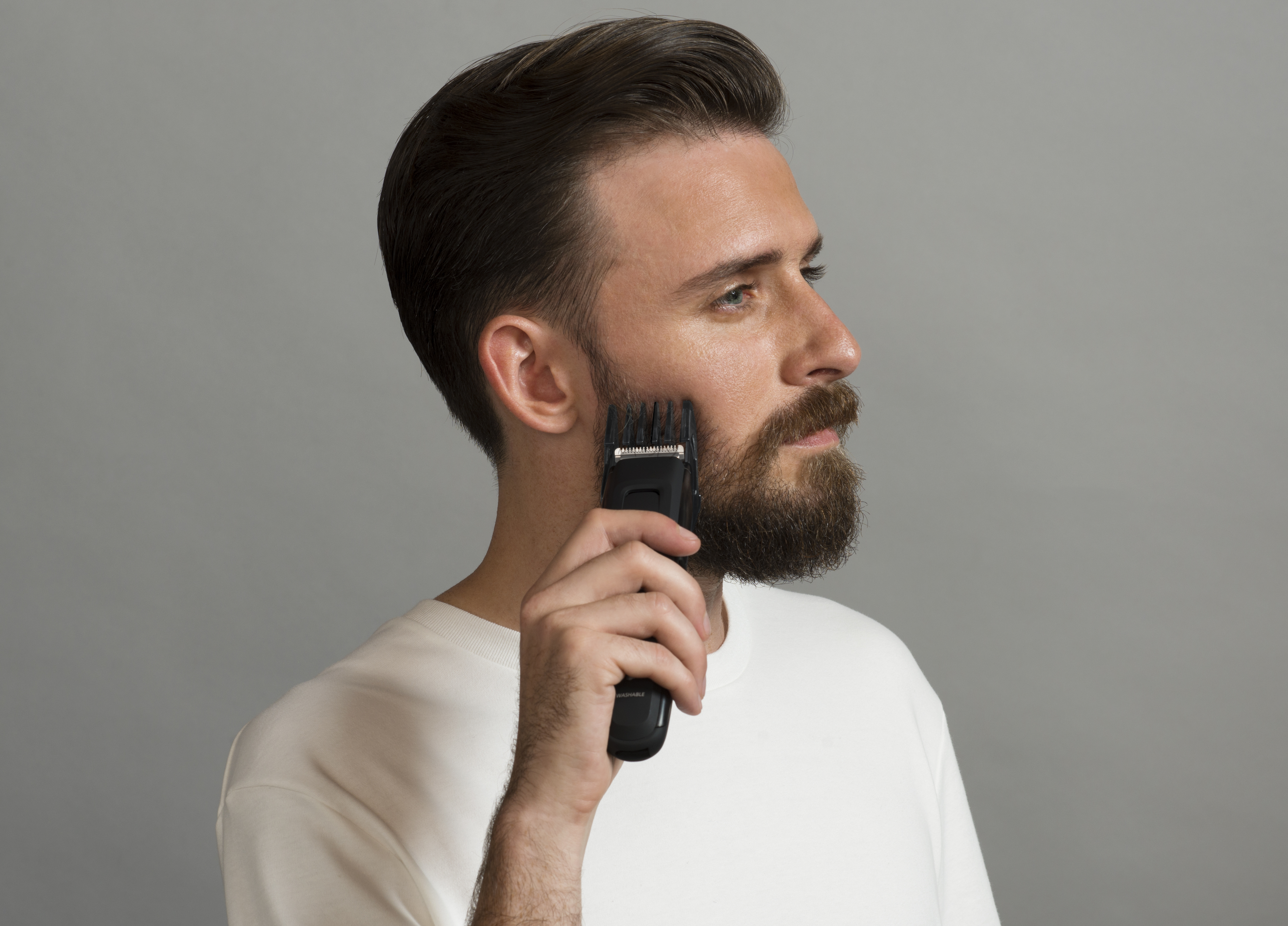 Uomo che taglia la barba con un trimmer nero. Profilo, camicia bianca, sfondo grigio.