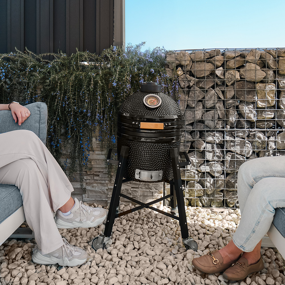 Zwei Personen in der Nähe eines schwarzen Grills mit Steinmauer und blauem Himmel.