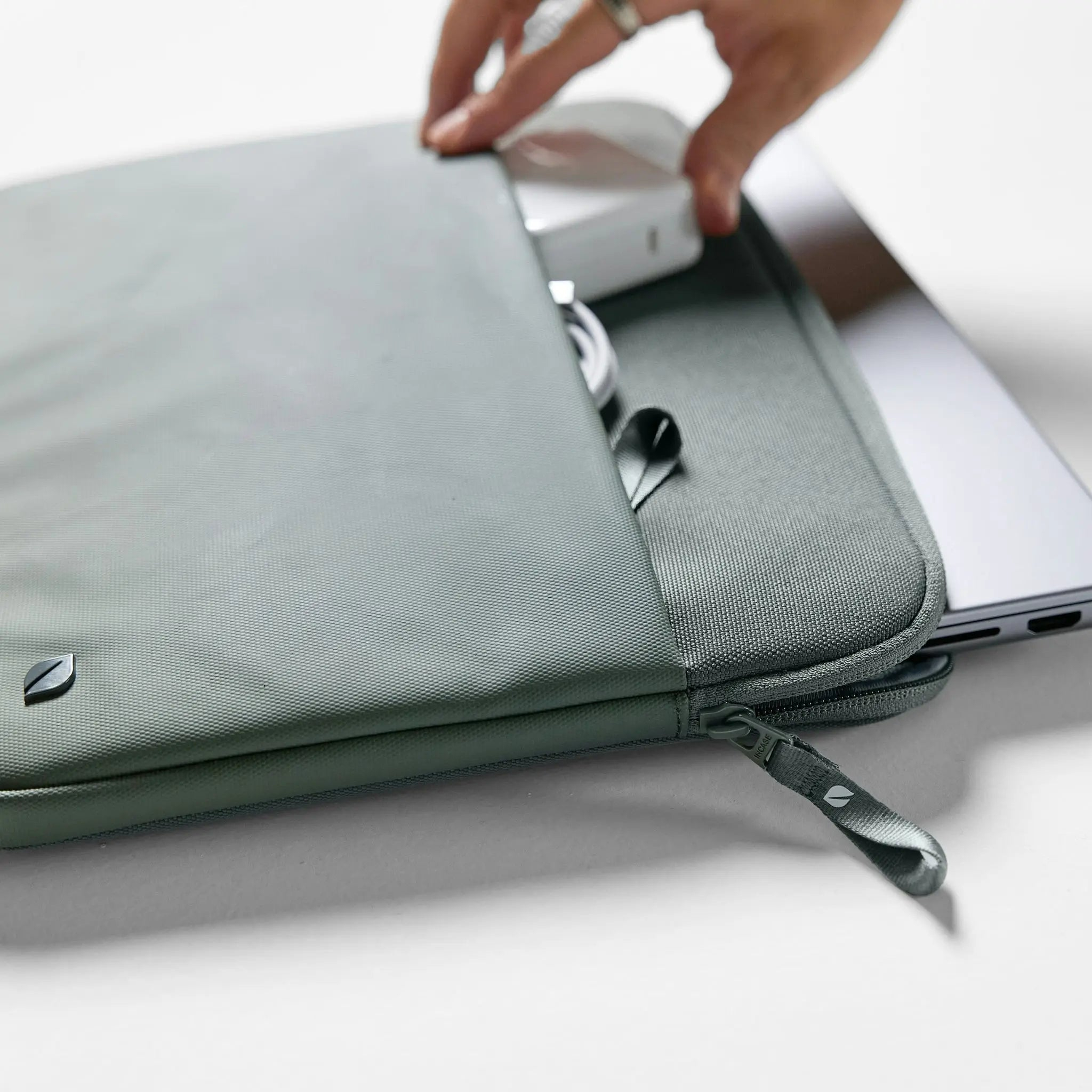 La mano di una persona apre una custodia grigia per laptop, rivelando un laptop e un caricatore.