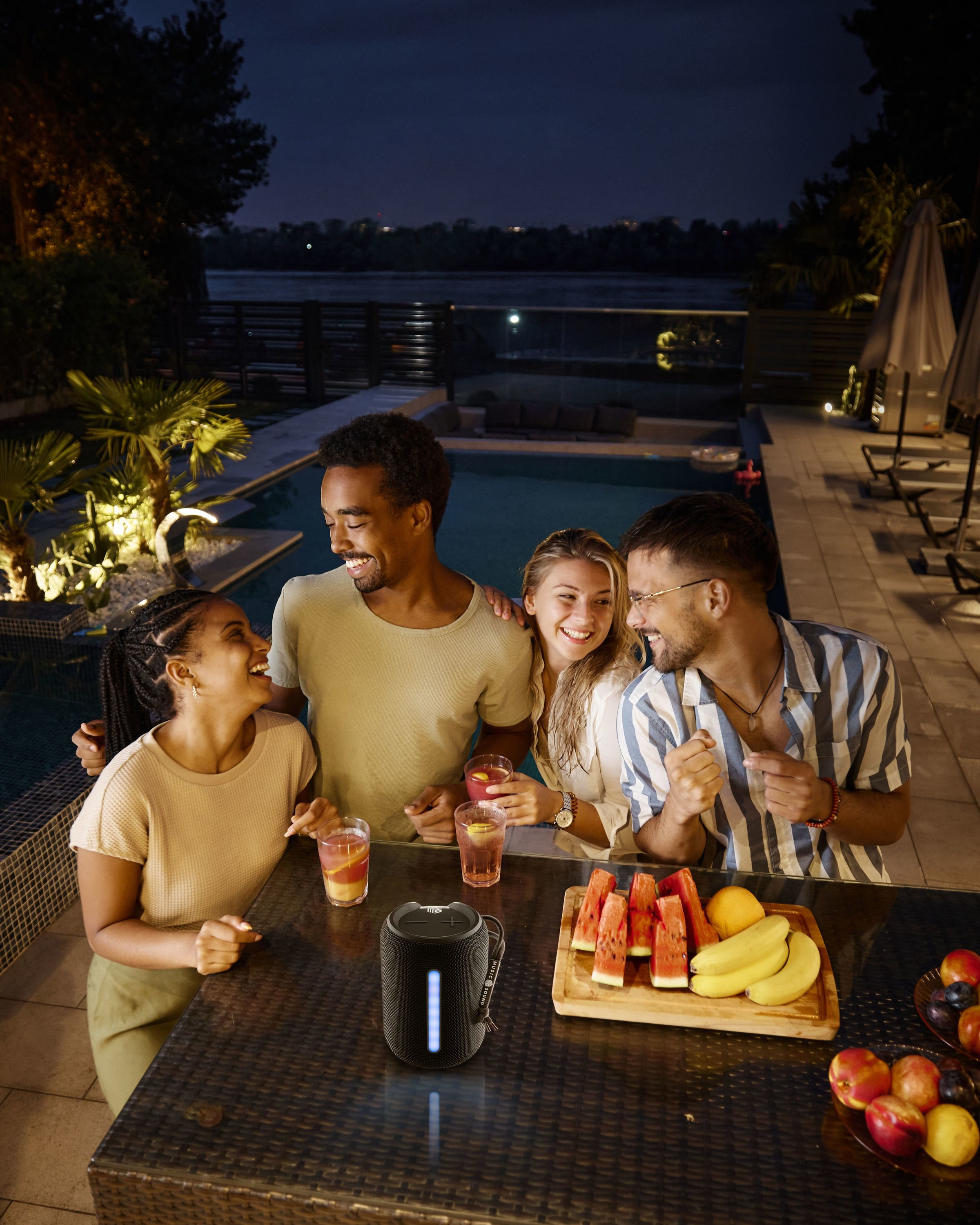 Grupa przyjaciół w nocy, przy basenie, pijąca koktajle, z głośnikiem na stole.