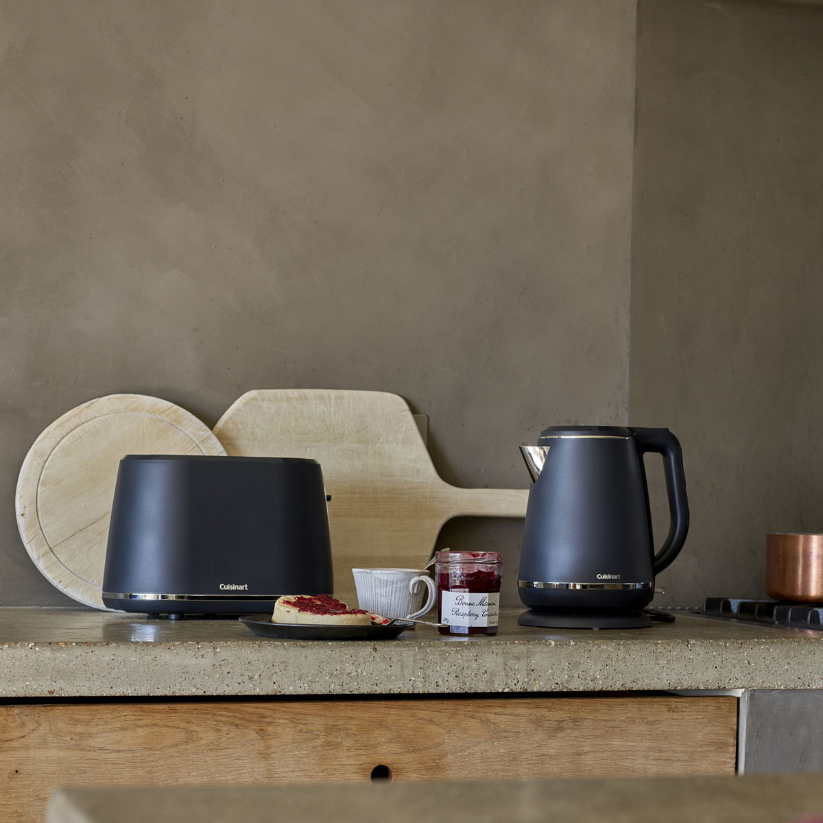 Scena kuchenna z tosterem, czajnikiem, deskami do krojenia, słoikiem dżemu i talerzem z jedzeniem.