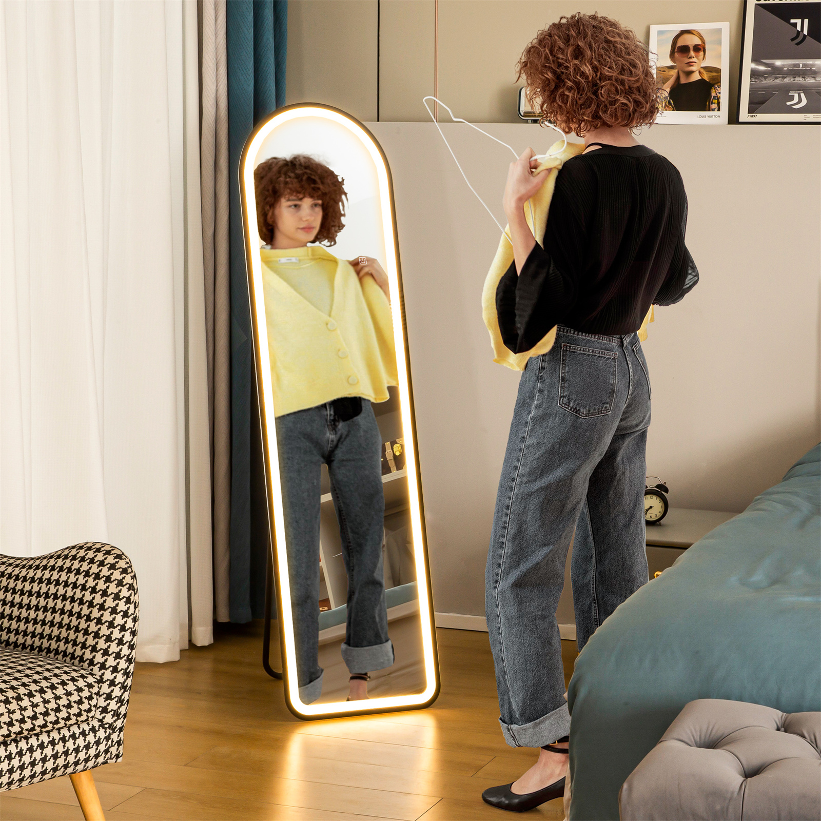 Frau steht in einem Zimmer vor einem Ganzkörperspiegel mit beleuchtetem Rahmen, Bett und Stuhl.