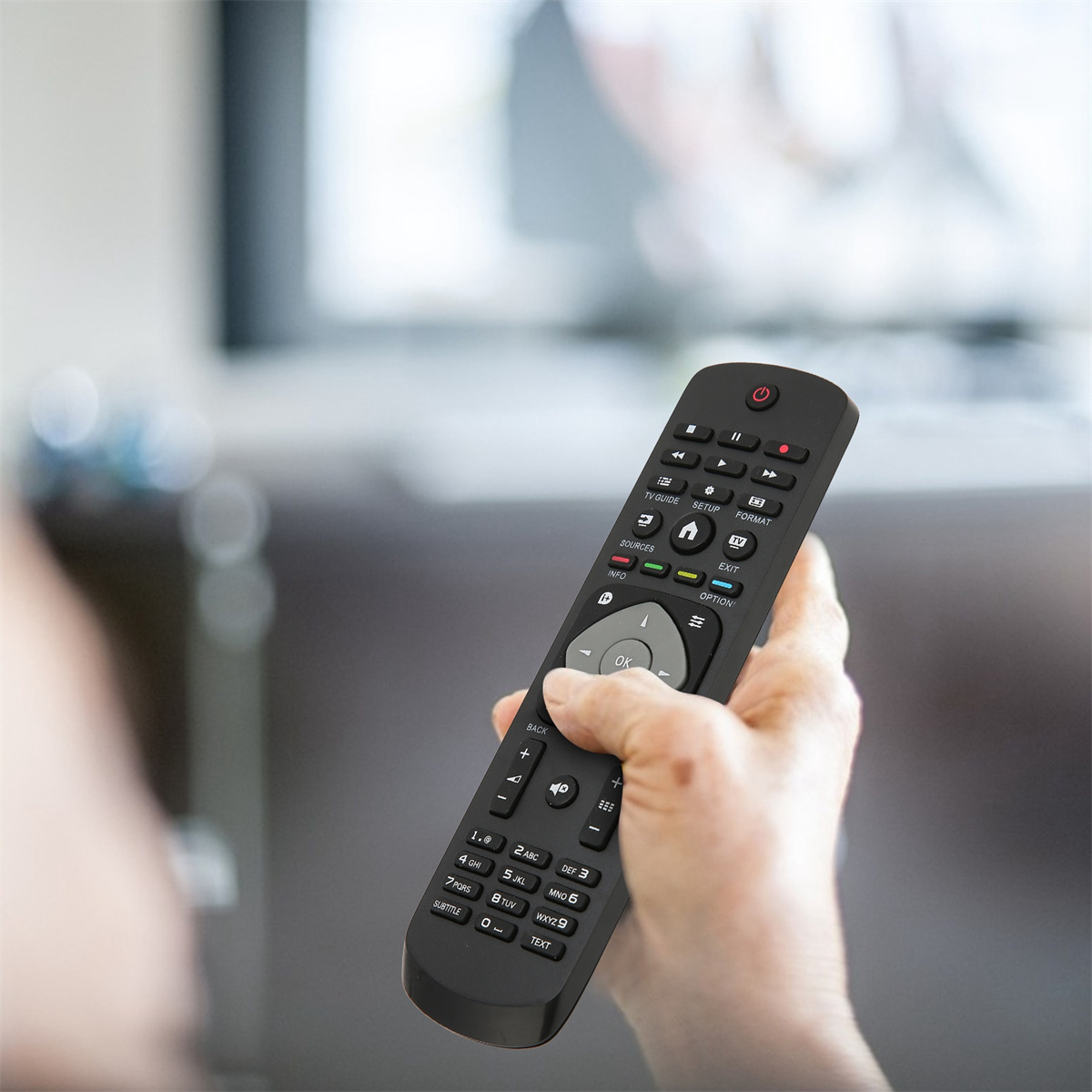 Die Hand einer Person, die eine schwarze TV-Fernbedienung hält, vor einem unscharfen Fernsehbildschirm.
