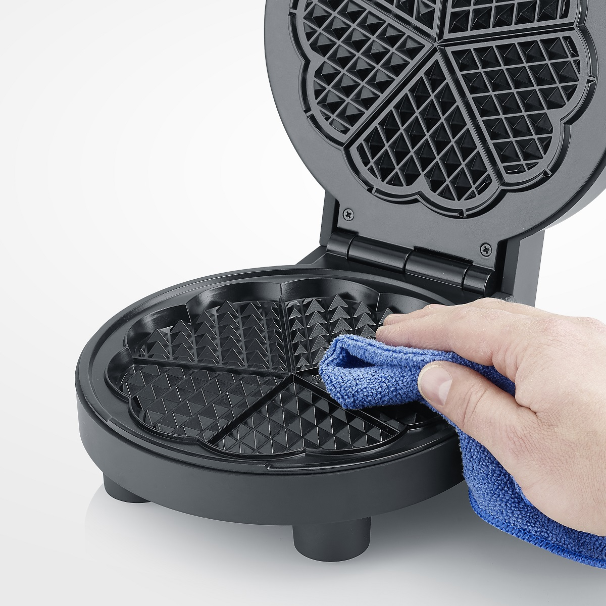 Hand reinigt Waffeleisen mit blauem Tuch, geöffnet auf weißer Oberfläche.
