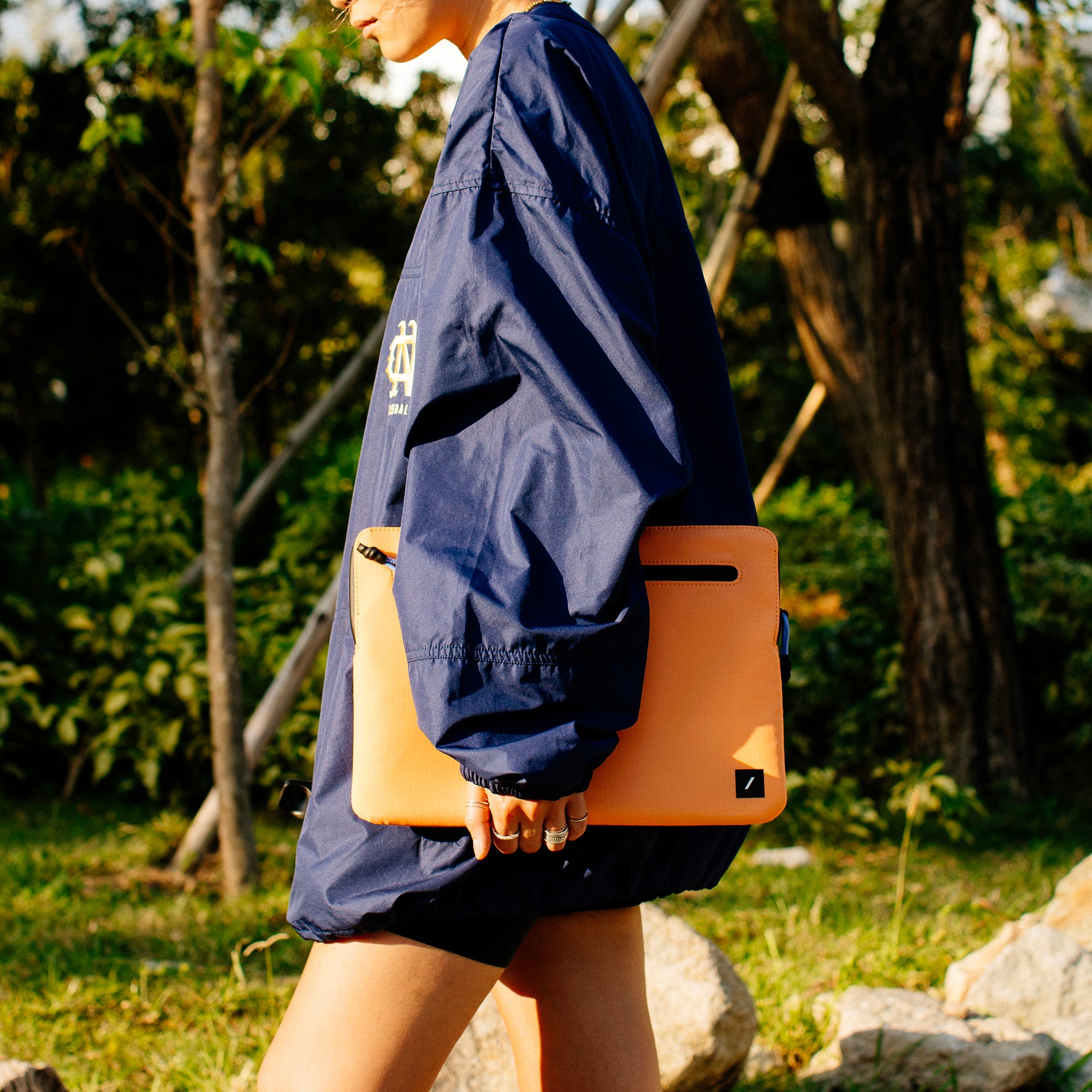 Person geht mit orangefarbener Laptop-Hülle, trägt blaue Jacke und Shorts.