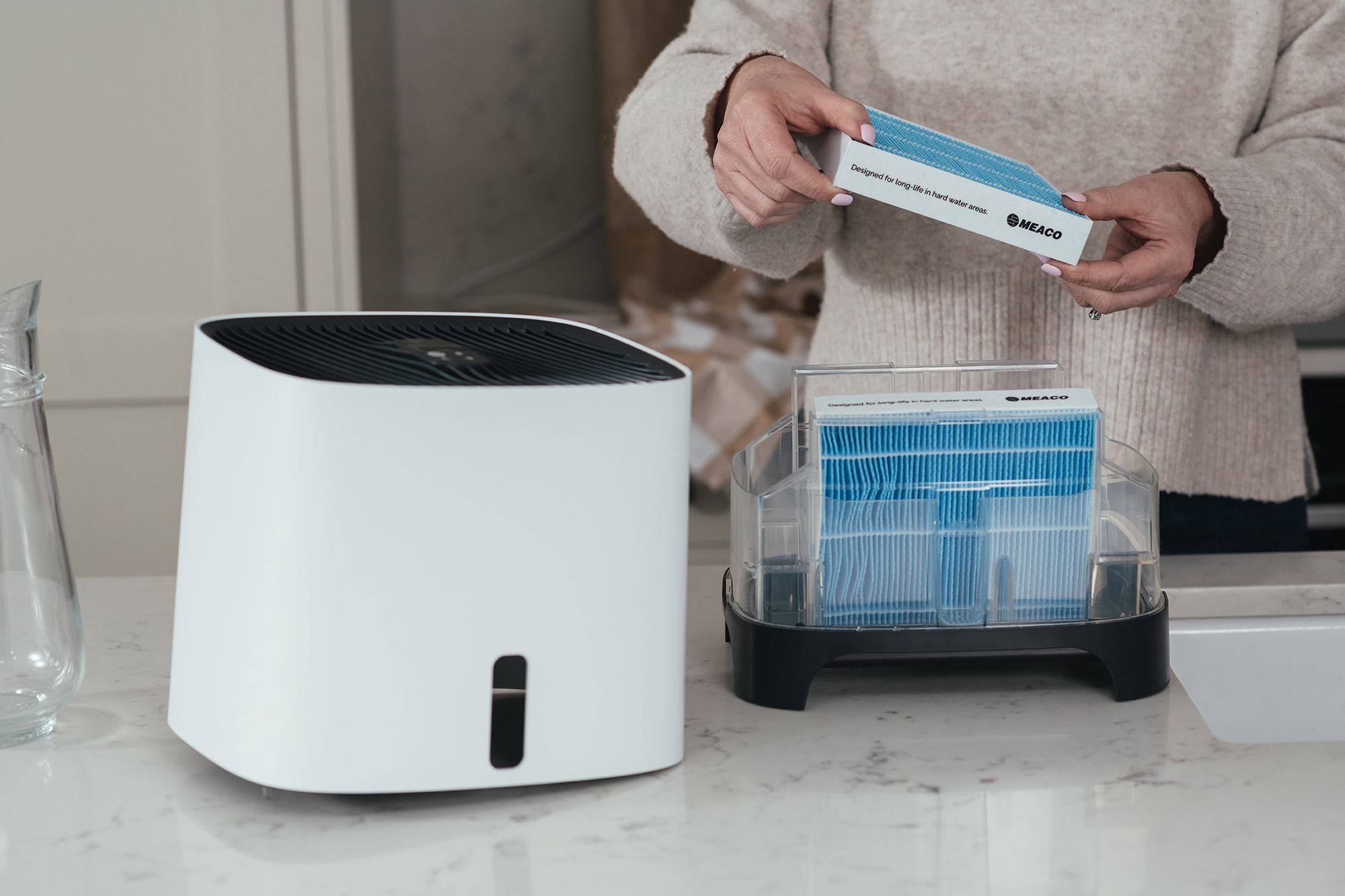 Osoba trzymająca opakowania filtrów w pobliżu białego oczyszczacza powietrza na blacie.