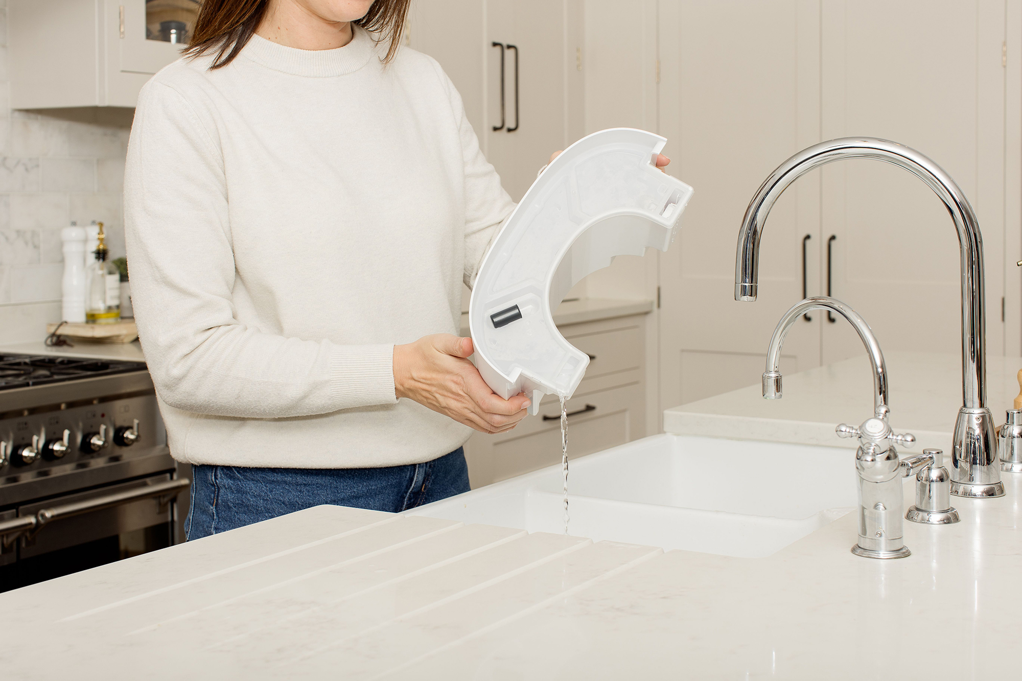 Kobieta trzyma biały plastikowy przedmiot nad zlewem, wypływa woda. Kran kuchenny po prawej stronie.