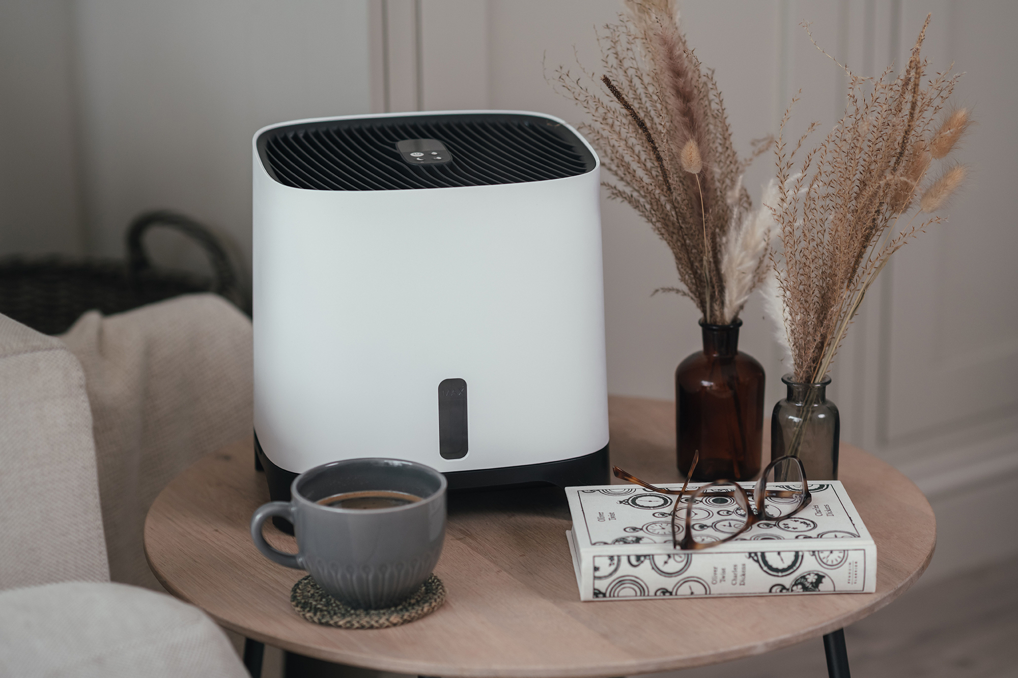 Biały oczyszczacz powietrza na stole z kawą, kwiatami i książką.