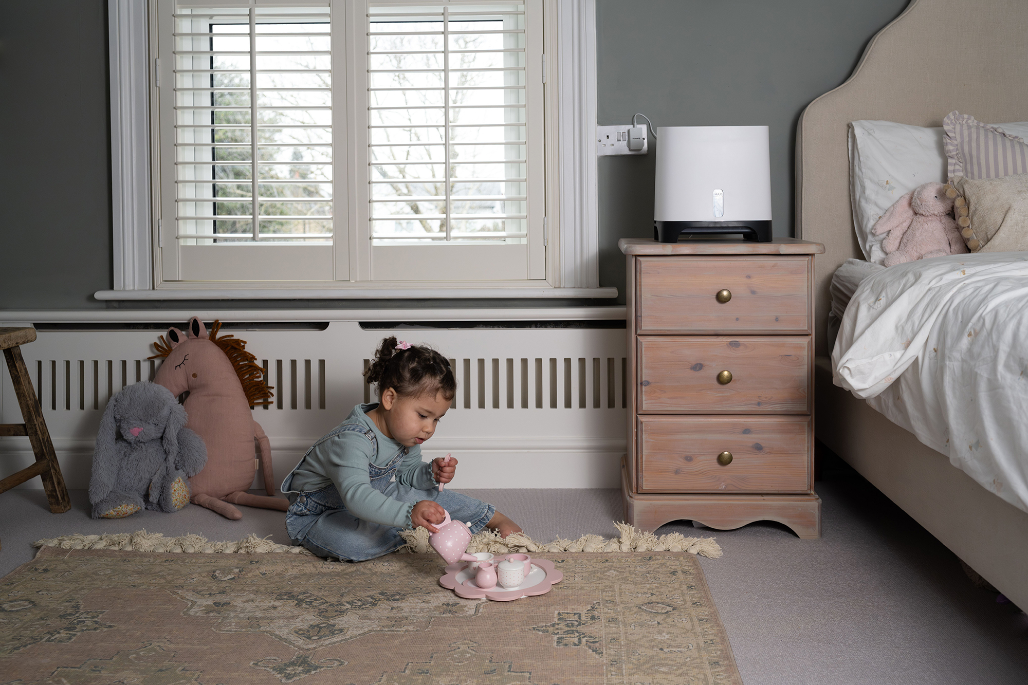 Mała dziewczynka bawiąca się zestawem do herbaty w pokoju. Widoczne łóżko, zabawki i nawilżacz.