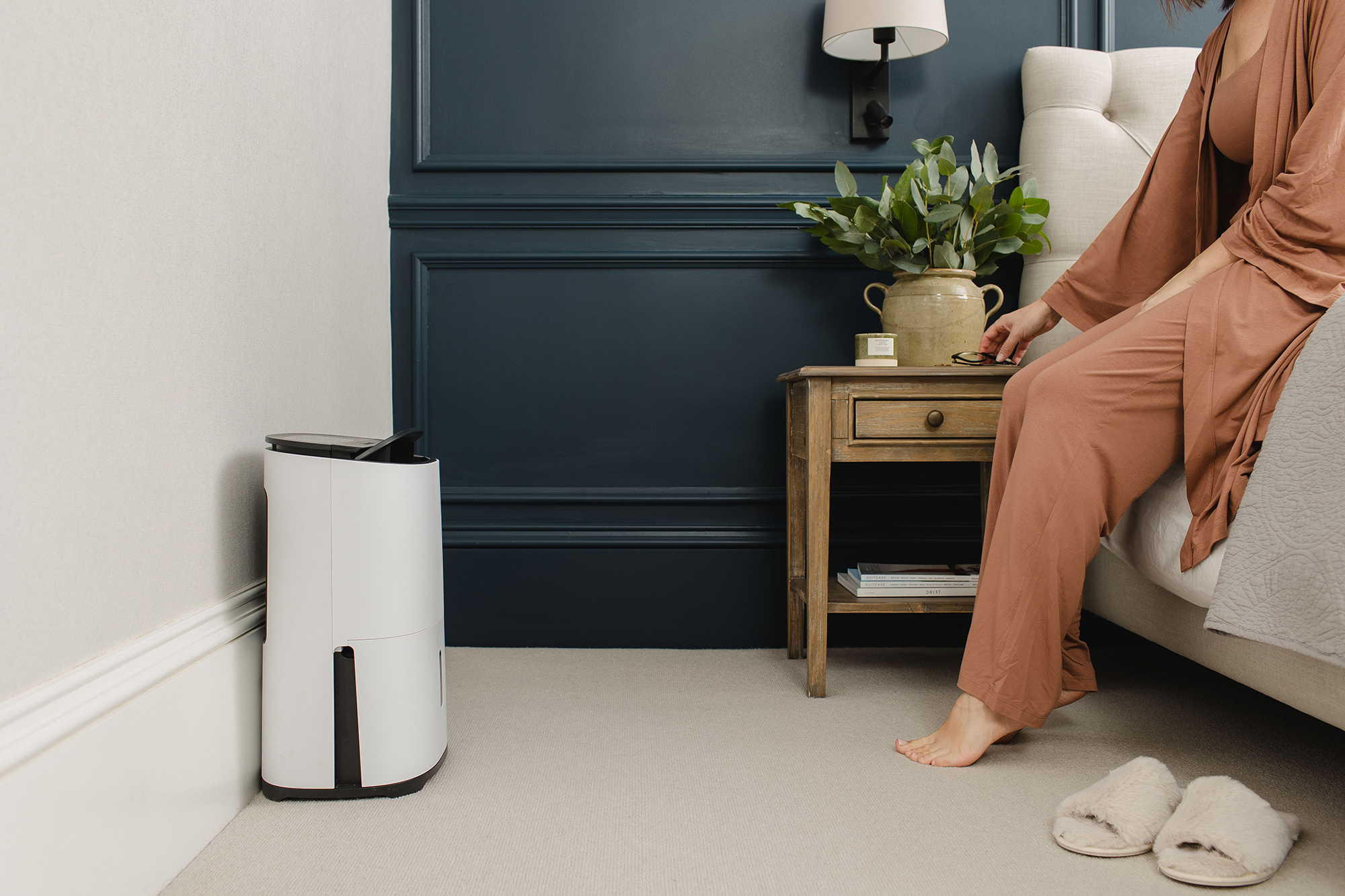 Kobieta w sypialni siedzi na łóżku. Nawilżacz stoi obok stołu.