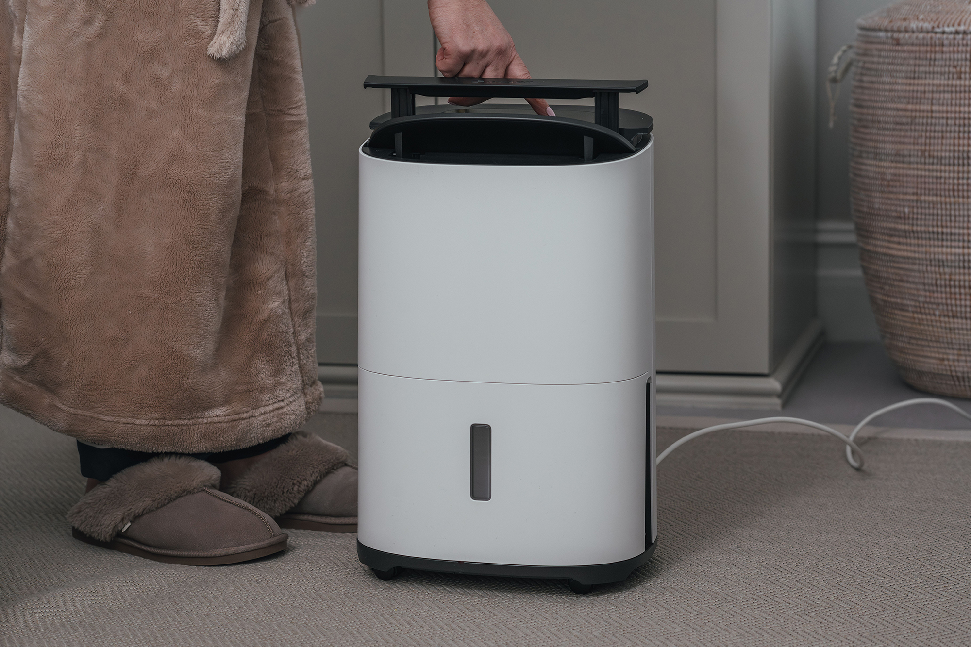 Biały osuszacz powietrza. Osoba w szlafroku i kapciach trzyma go.