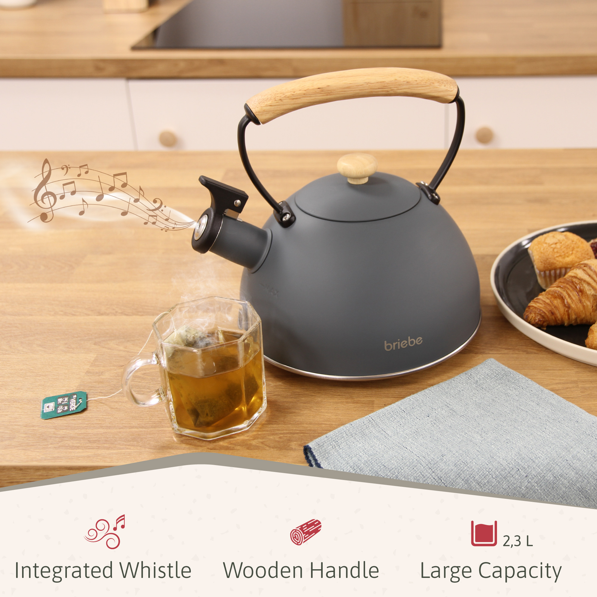 Ein grauer Wasserkessel mit Holzgriff steht neben einer Tasse Tee, einem Gebäckteller und einem Leinentuch.