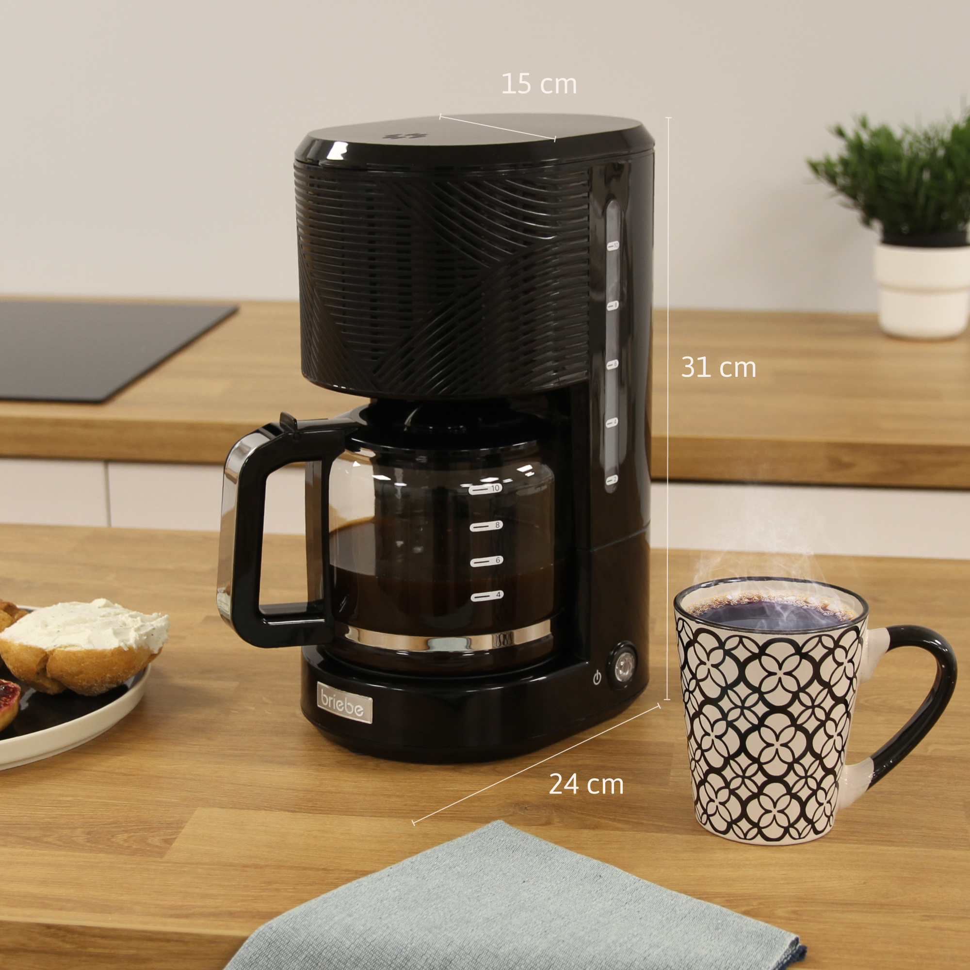 Una cafetera negra con una jarra de vidrio, una taza y dimensiones sobre la encimera.