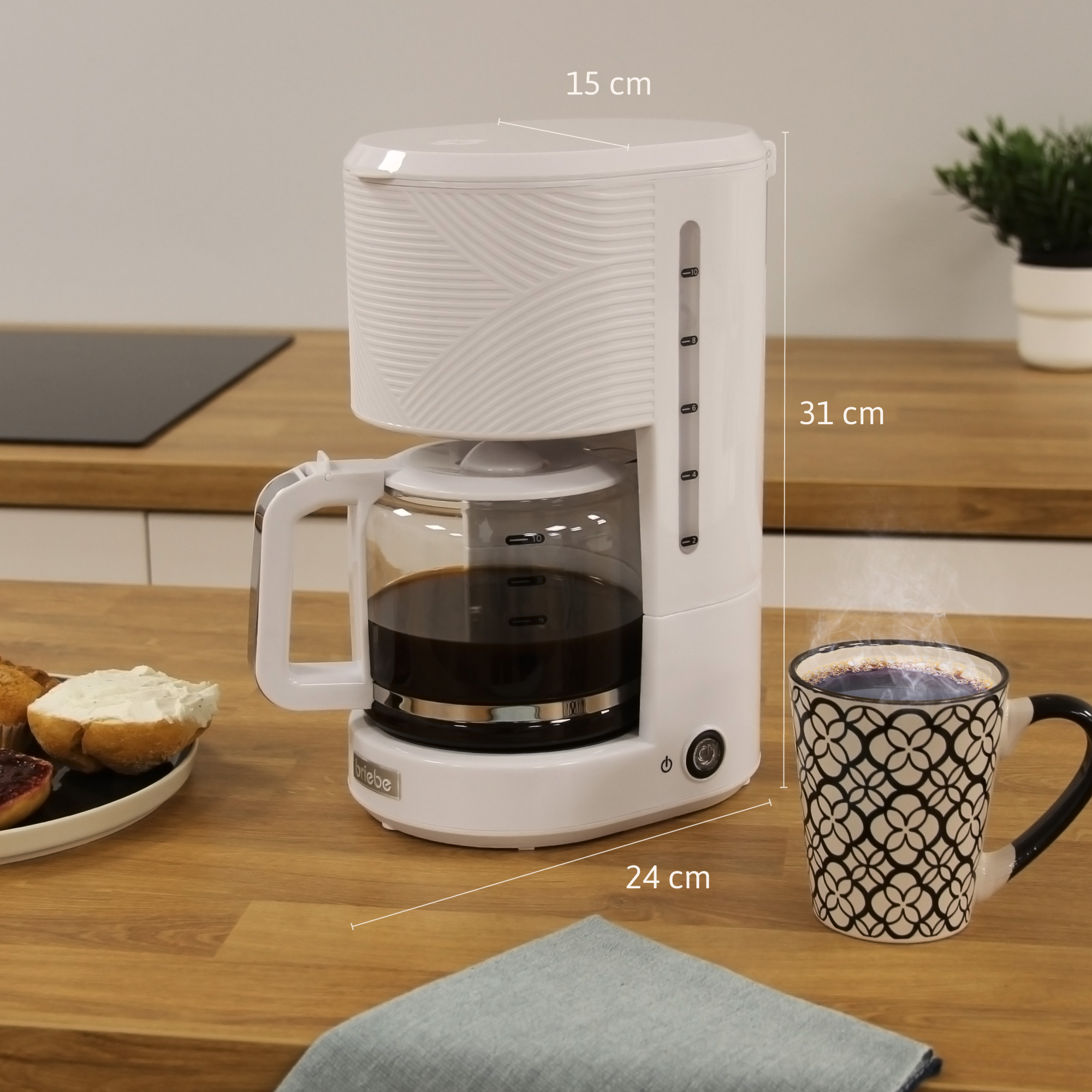 Una cafetera blanca con una jarra de cristal, una taza y una servilleta sobre una mesa de madera.