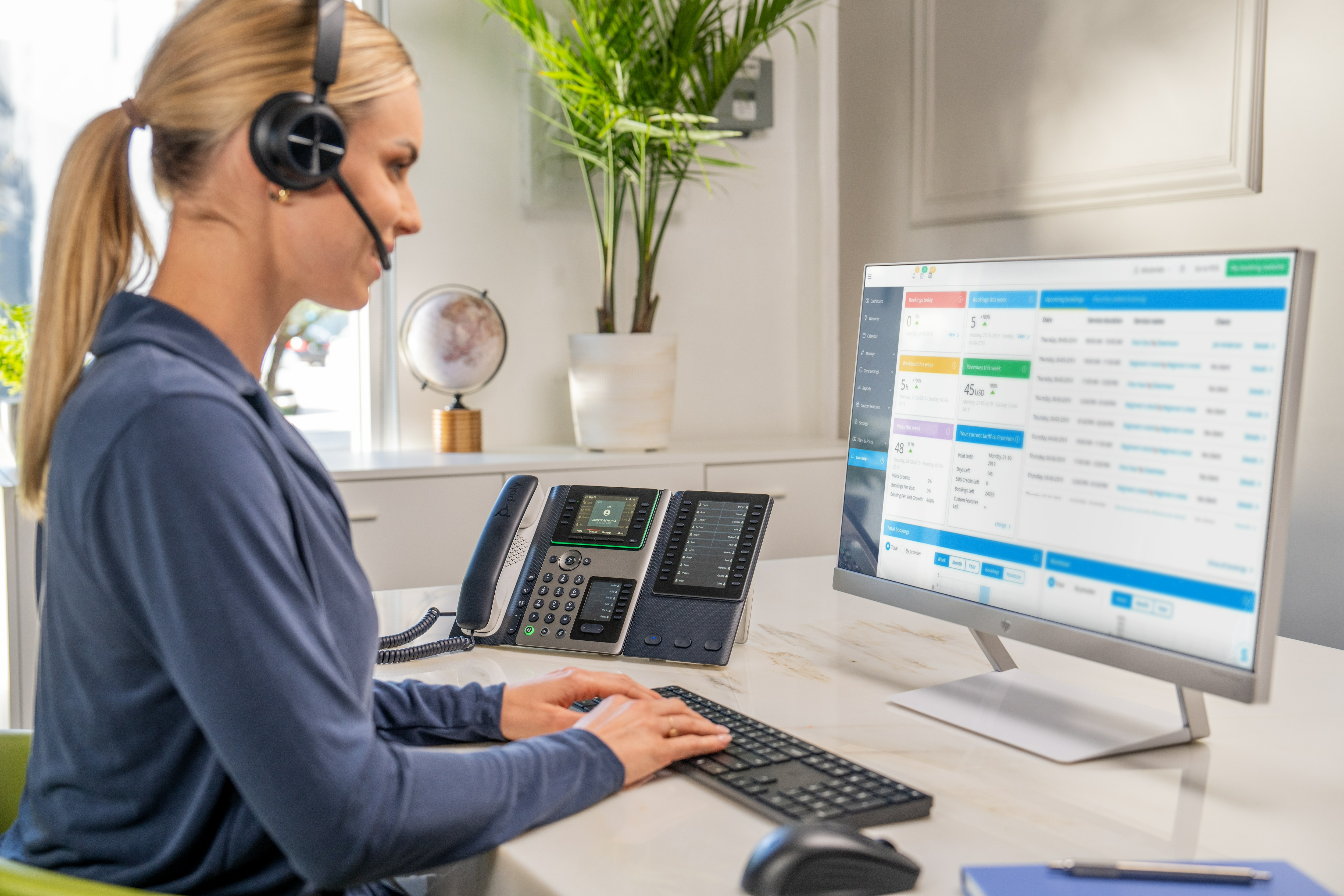 Una donna con auricolare lavora a una scrivania con un computer e due telefoni.