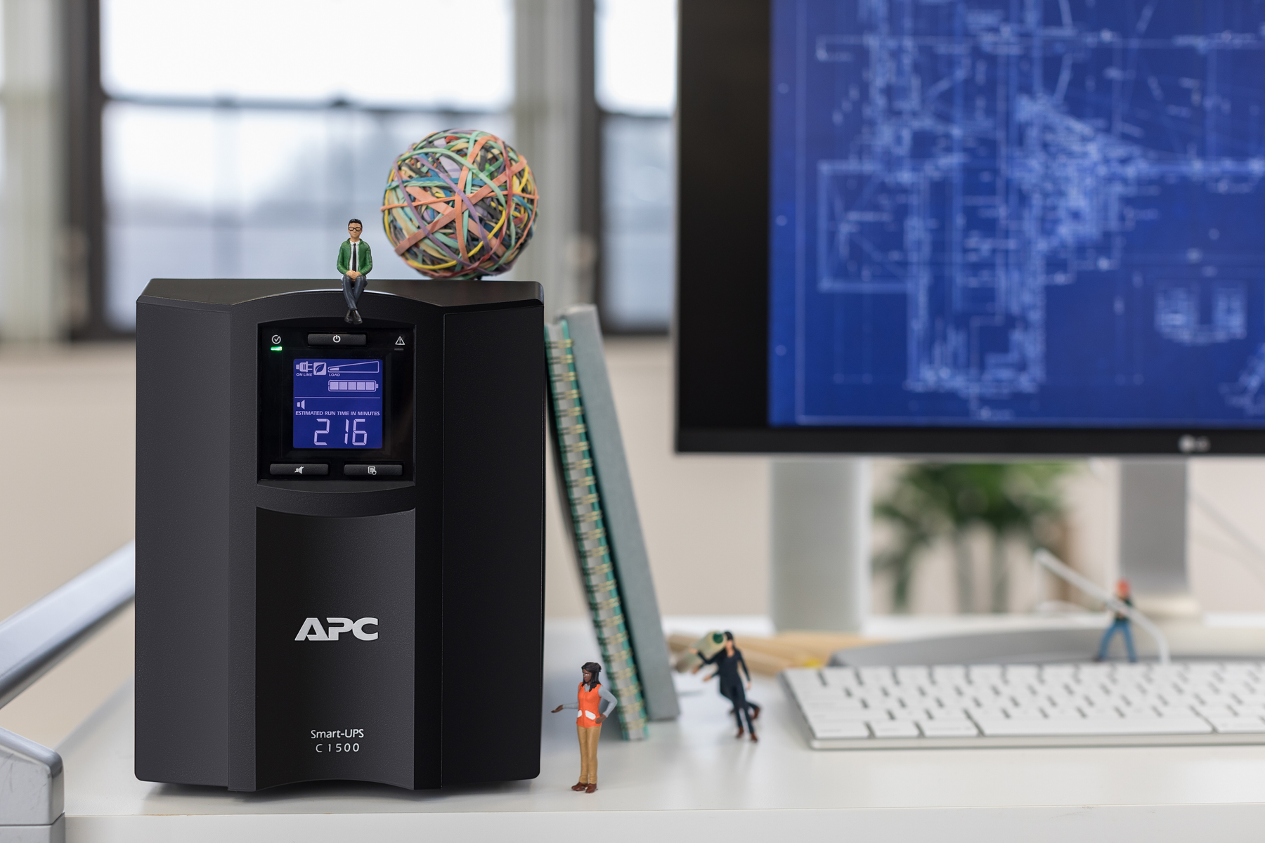 Eine APC Smart-USV auf einem Schreibtisch mit Miniaturfiguren und einem Bauplan im Hintergrund.