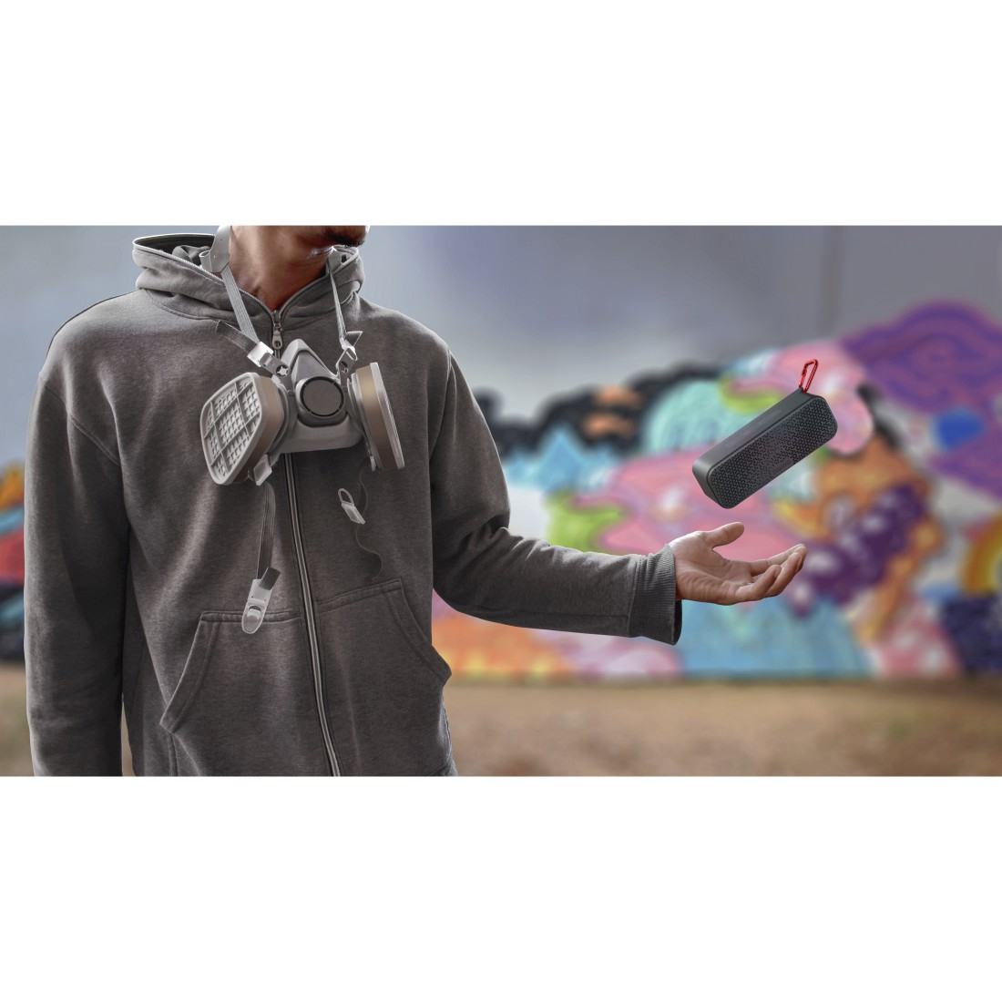 Mężczyzna w masce gazowej trzyma czarny głośnik, graffiti w tle.