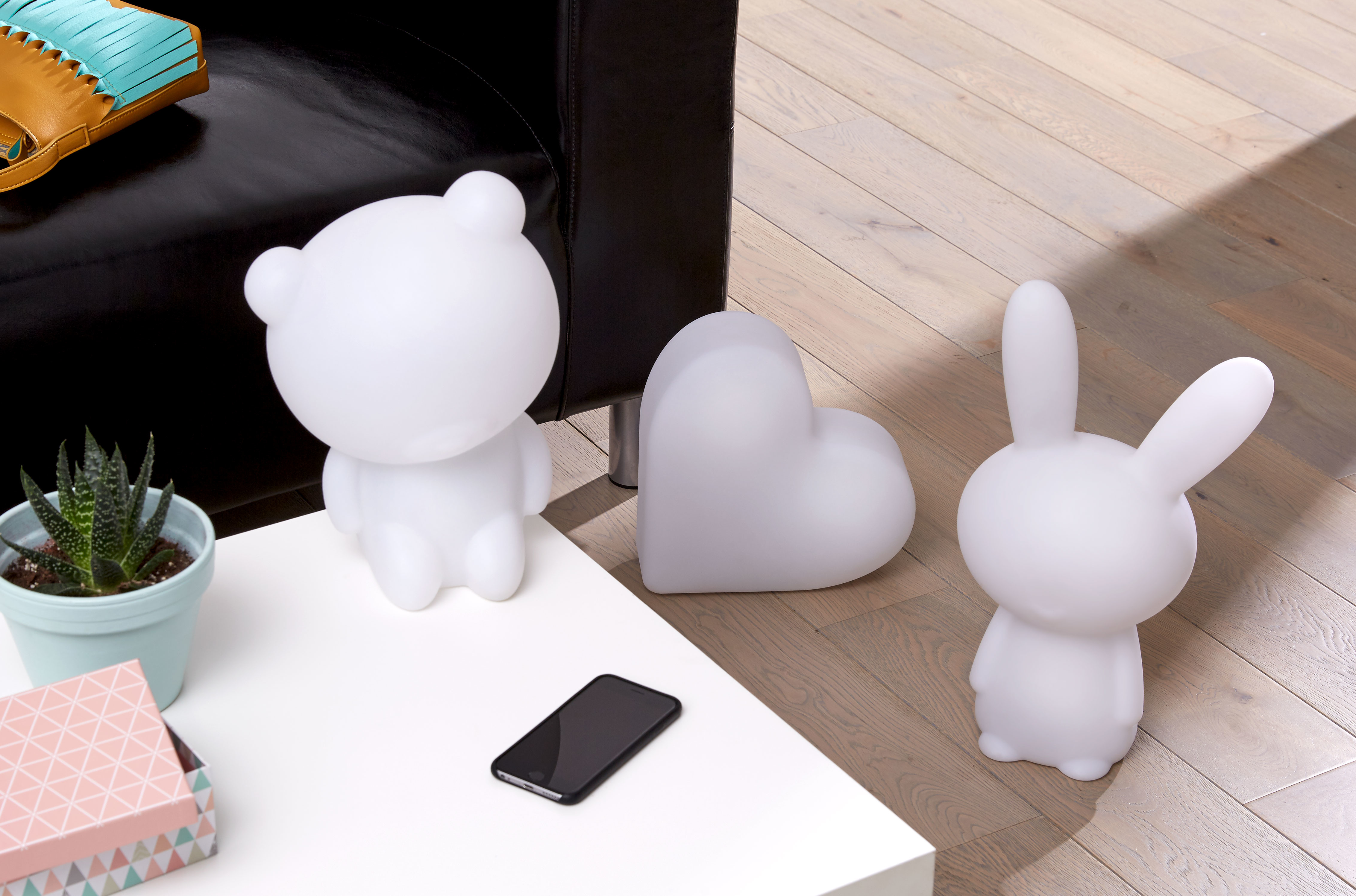 Białe lampki w kształcie misia, serca i królika na stole. Na stole również telefon i roślina. Drewniana podłoga.