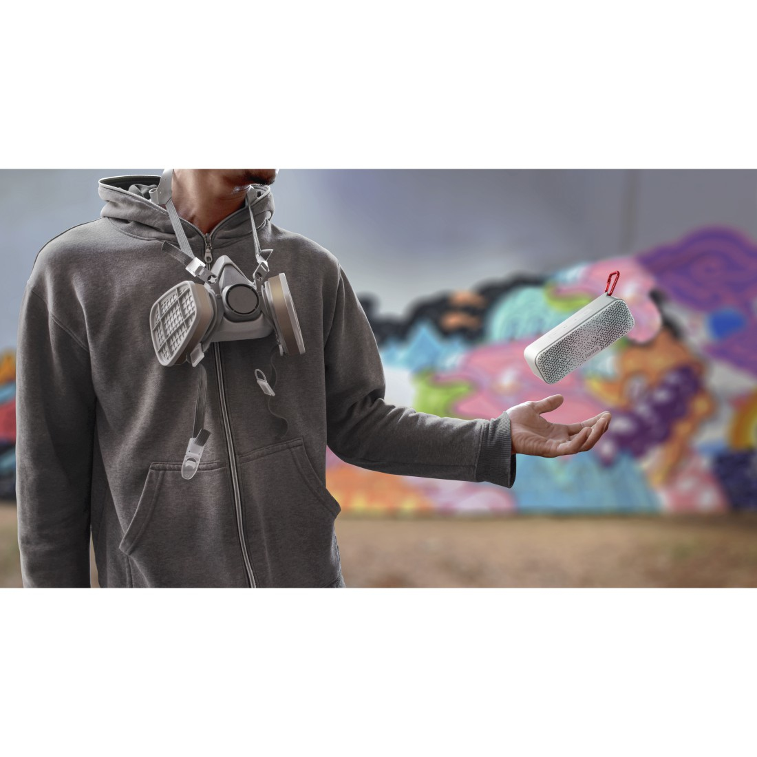 Mężczyzna w szarej bluzie z kapturem trzyma mały biały głośnik. Maska gazowa wisi na szyi. Tłem jest mural.