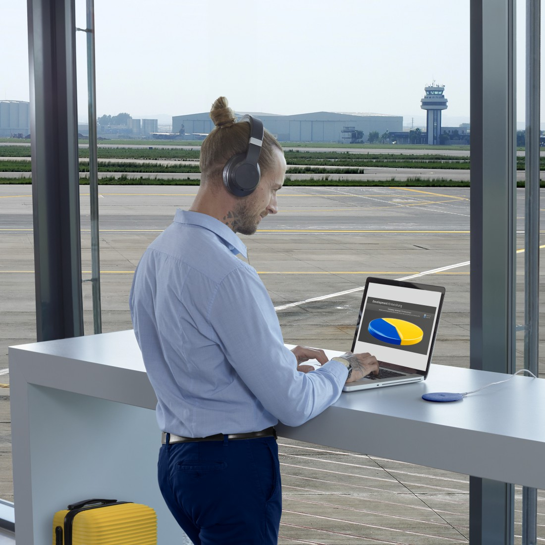 Mężczyzna na lotnisku z laptopem, wykres kołowy na ekranie. Słuchawki na uszach, samolot i wieża kontroli w tle.