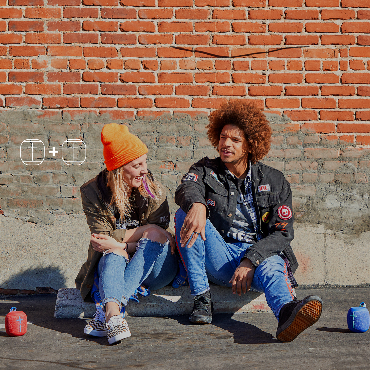 Dwie osoby siedzące przy ceglanej ścianie, z dwoma małymi głośnikami i innymi przedmiotami.