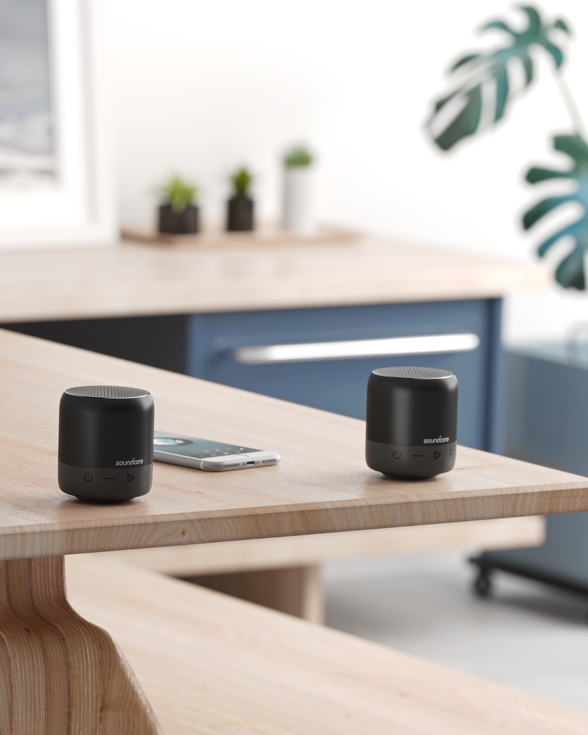 Dwa czarne głośniki Soundcore na drewnianym stole. Telefon między nimi. Rośliny w tle.