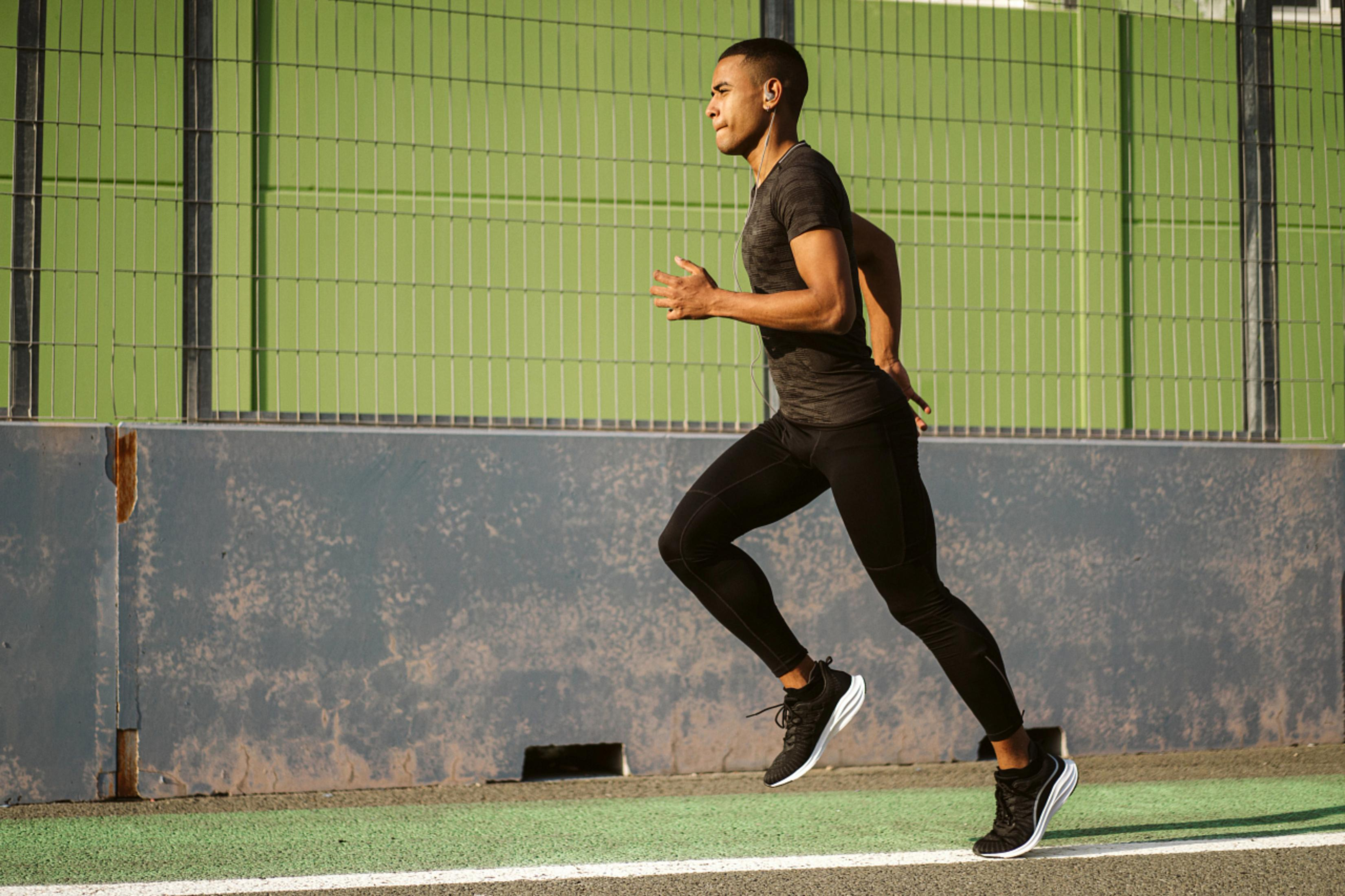 Un uomo in abbigliamento sportivo corre su una pista; maglietta scura e leggings, con uno sfondo sfocato di verde e grigio.