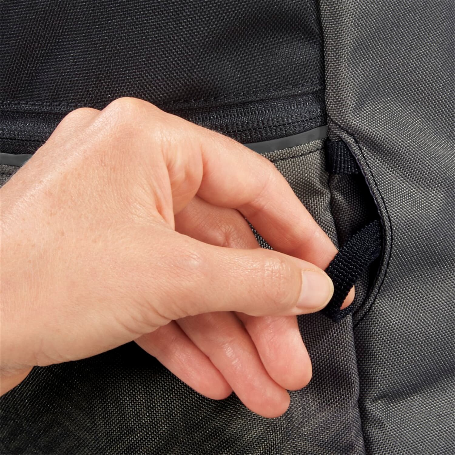 Eine Hand zieht eine schwarze Schlaufe an einer grauen Tasche.