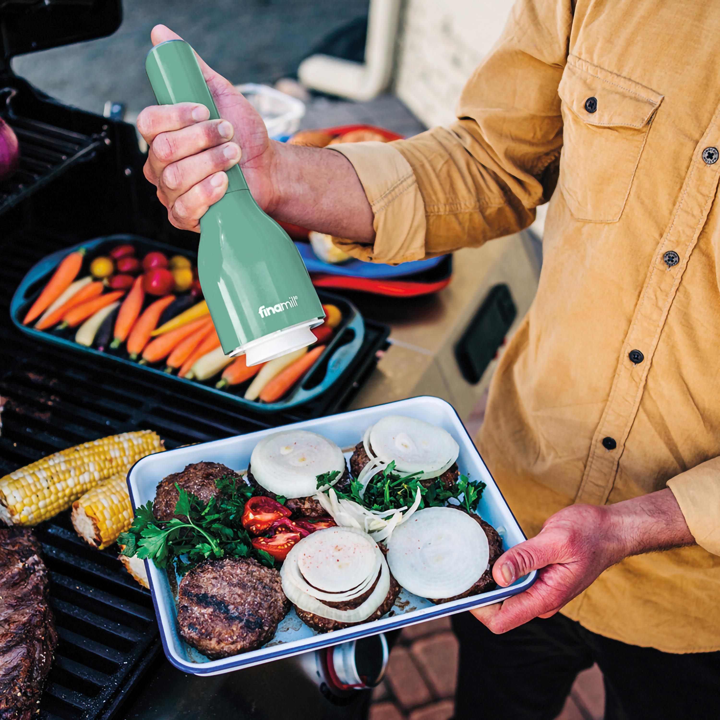 Person grillt Burger. Hält Fleischwolf und Tablett mit Burgern und Zwiebeln. Grill im Hintergrund.