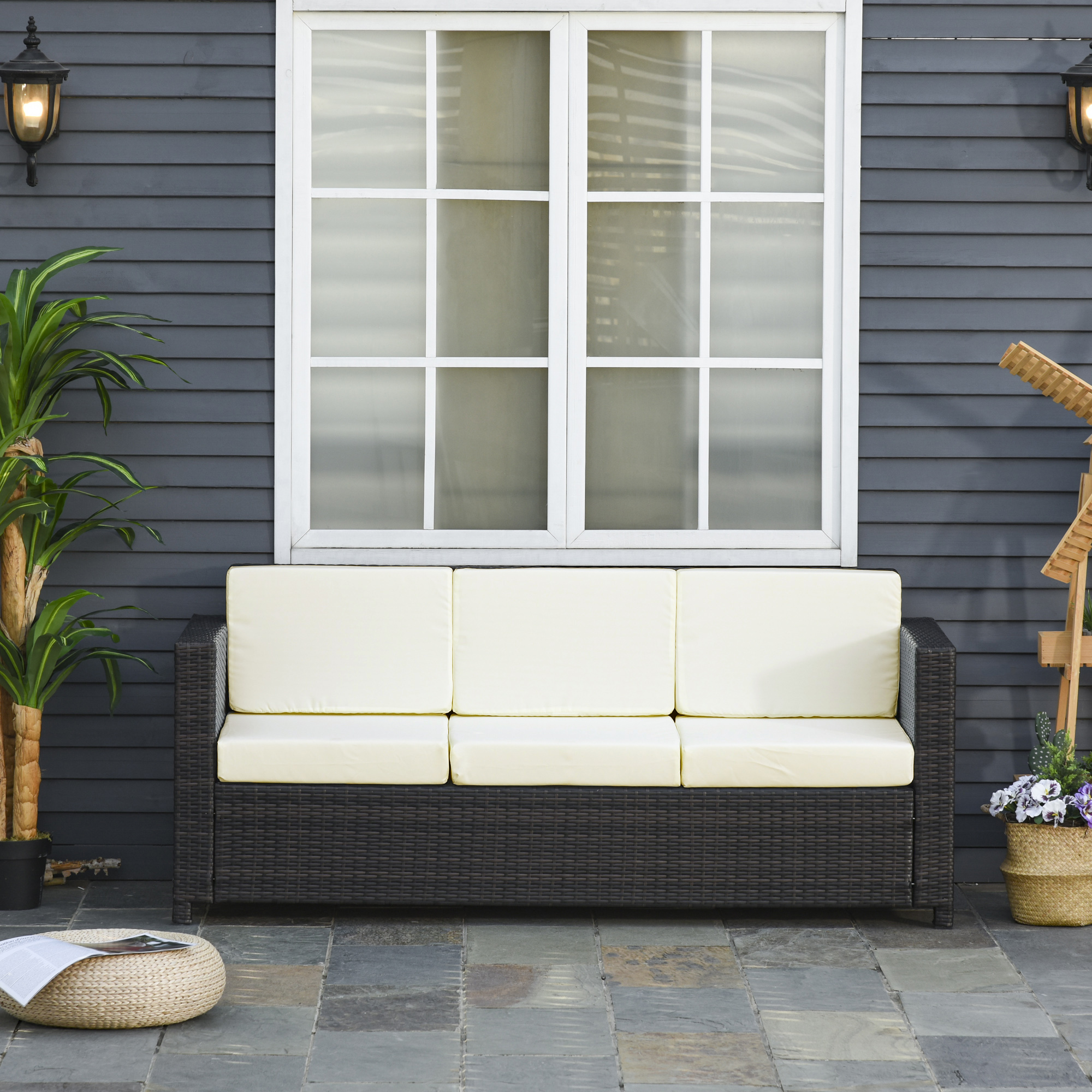 Geflochtenes Outdoor-Sofa mit cremefarbenen Kissen, vor einem Haus.