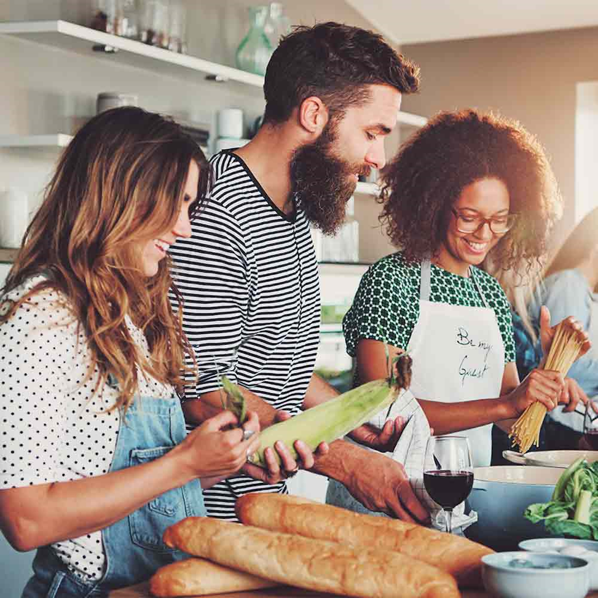 Drei Personen in einer Küche bereiten Essen zu. Einer hält Mais, ein anderer Nudeln, andere Wein und Brot.