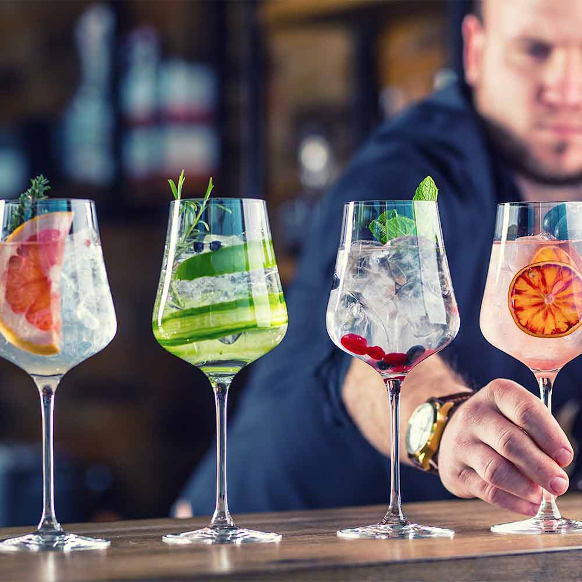 Vier Cocktailgläser mit verschiedenen Getränken. Barkeeper im Hintergrund, mit Uhr.