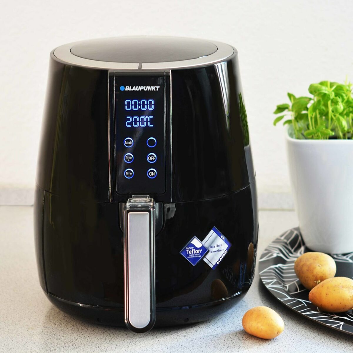 Zwarte airfryer met digitaal display. Aardappelen en een plantenpot op de achtergrond.