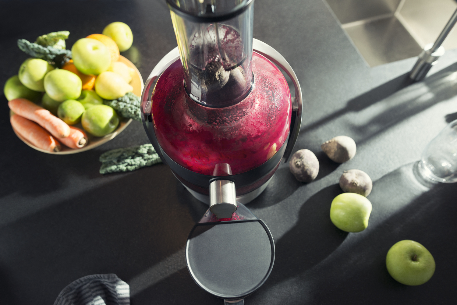 Un estrattore di succo con succo rosso, accanto a frutta e verdura su un tavolo scuro.
