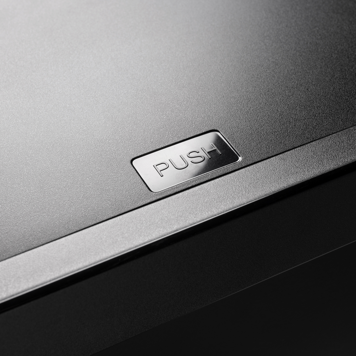 Close-up van een zwart oppervlak met een zilveren 'PUSH'-knop. Het oppervlak lijkt gemaakt van een textuurrijk materiaal.