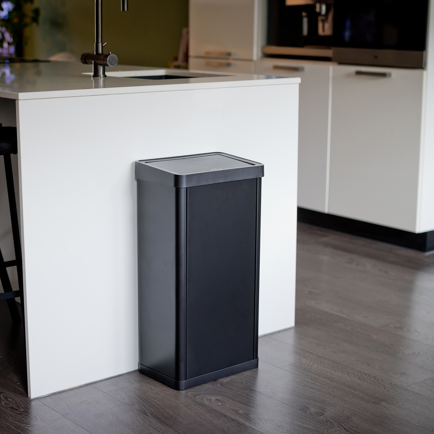 Zwarte rechthoekige vuilnisbak naast een witte keukenkast. Houten vloer.