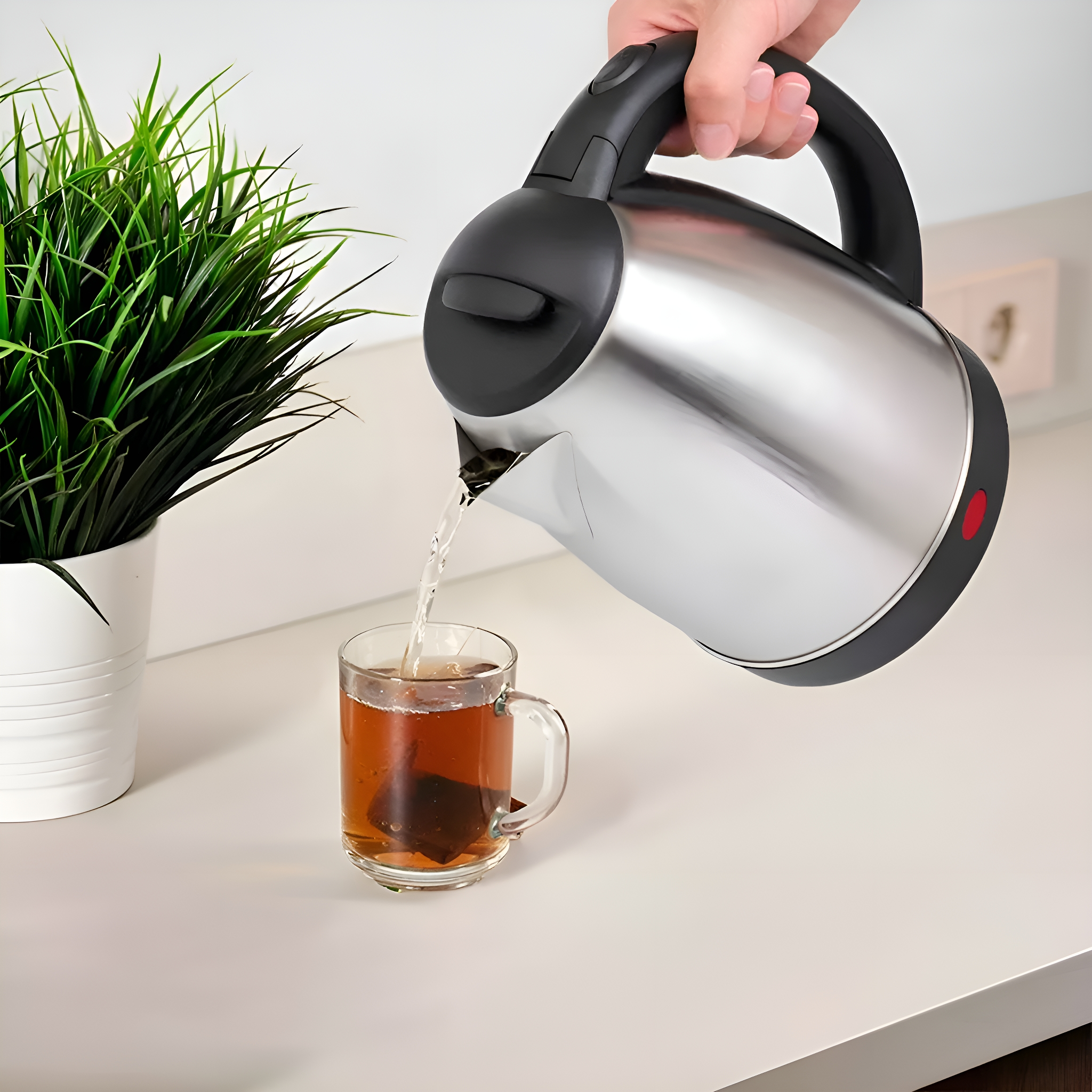 Edelstahlkessel gießt Tee in eine Glas-Tasse mit Teebeutel auf einer weißen Oberfläche.
