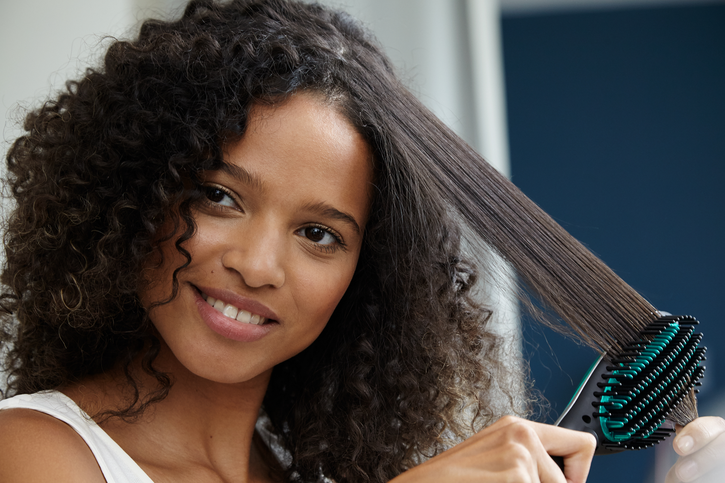 A woman uses a hair straightener on her curly hair, smiling. The background is a bathroom.