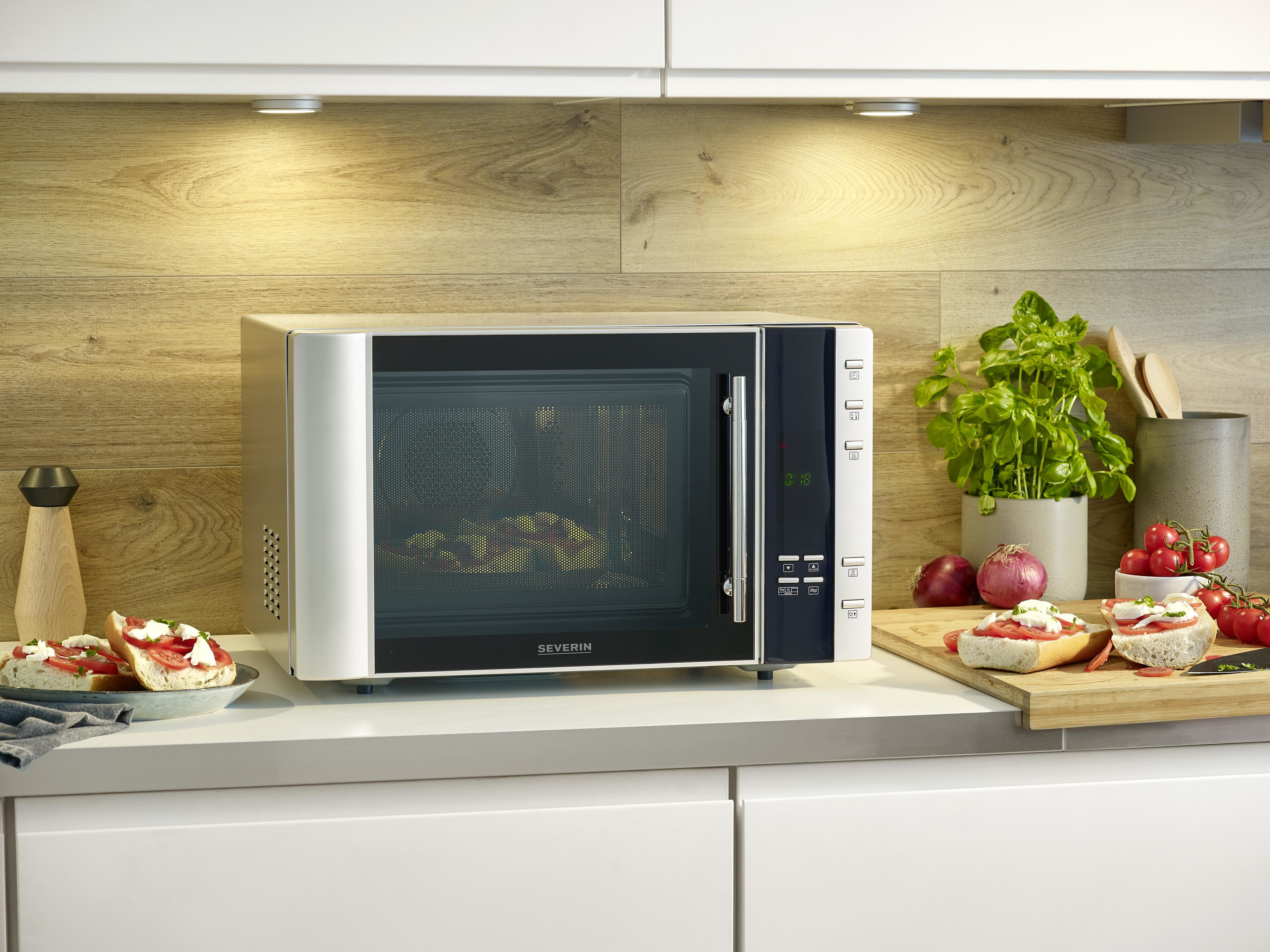 Kuchenka mikrofalowa stoi na białym blacie z jedzeniem wokół, w otoczeniu kuchni.