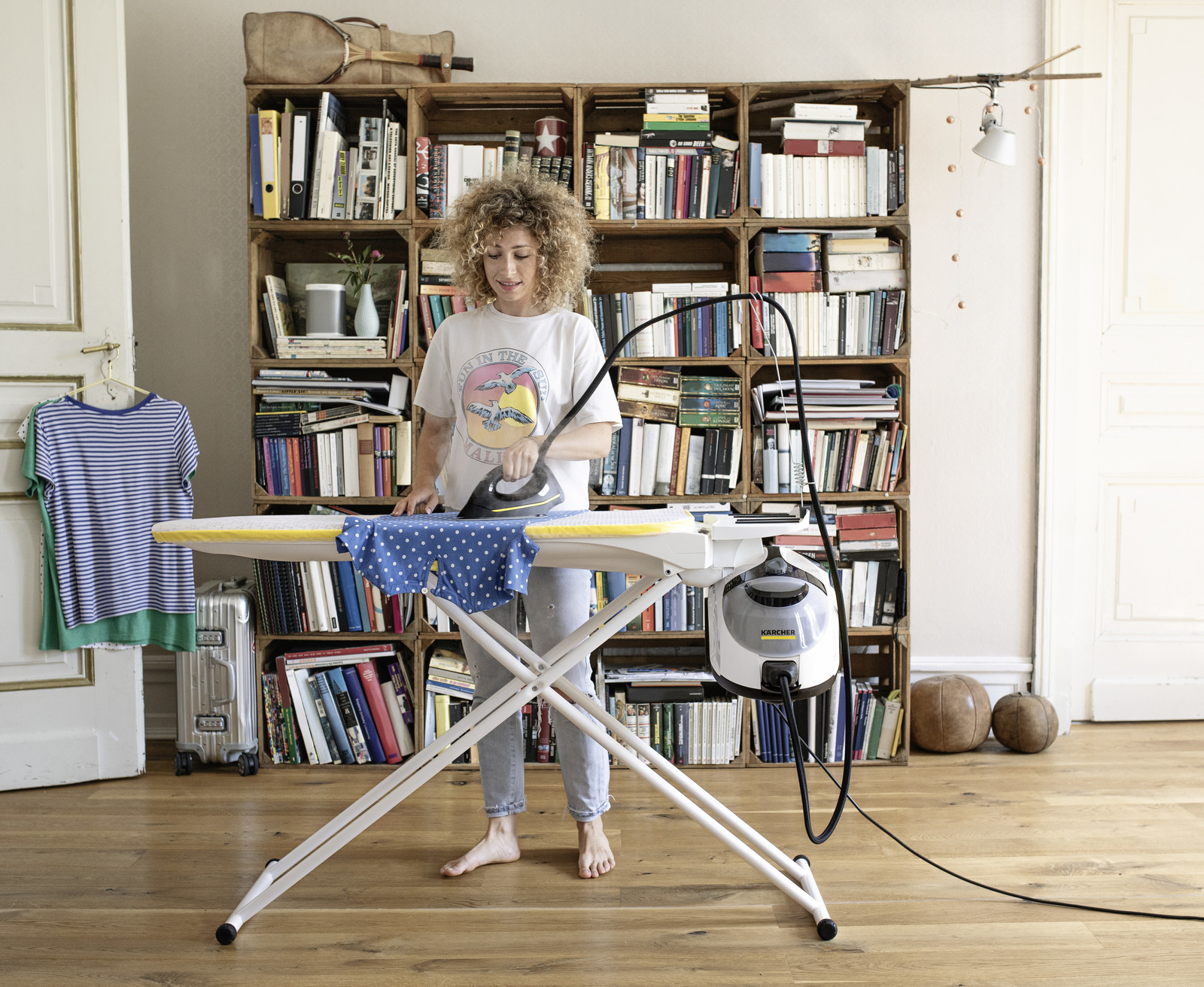 Kobieta prasująca niebieską koszulę w grochy na desce do prasowania w dobrze oświetlonym pokoju z książkami.
