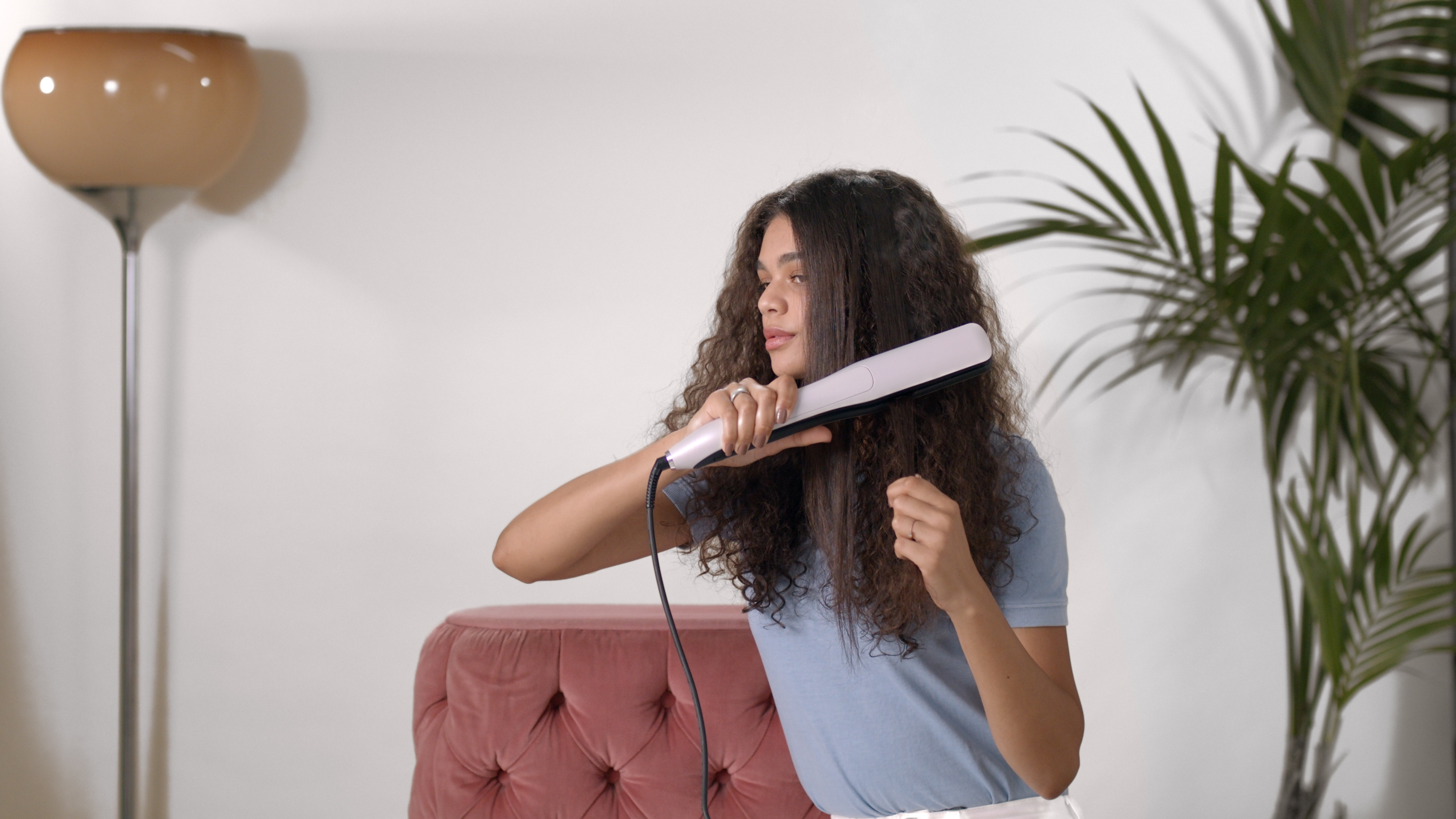 Vrouw die een stijltang gebruikt. Krullend haar en een roze stoel op de achtergrond.