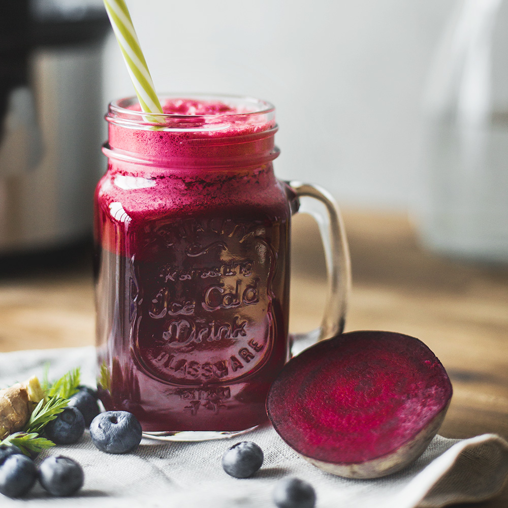 Ein Glas roter Saft mit Strohhalm, Blaubeeren und einer halbierten Bete auf einem Holztisch.