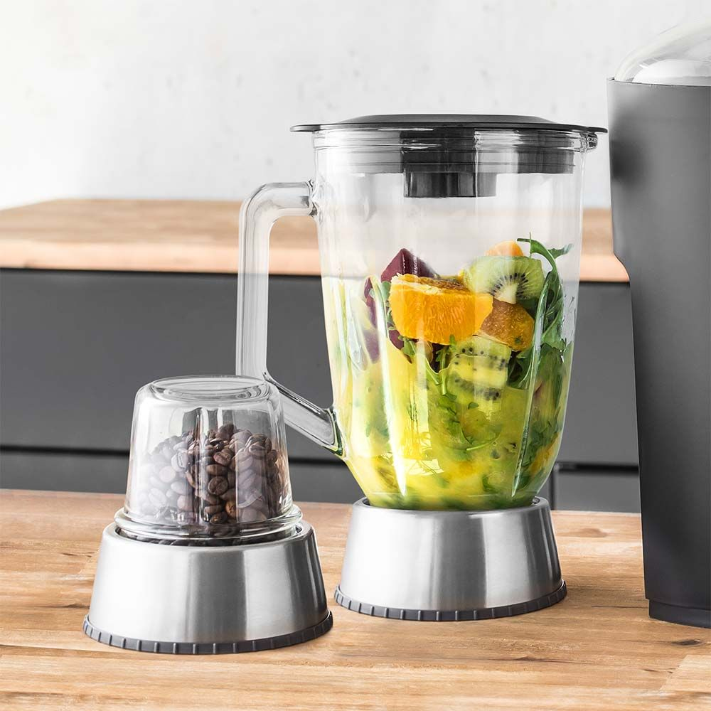 Mixer mit Obst und Gemüse, ein kleiner Behälter mit Kaffeebohnen, auf einem Tisch.