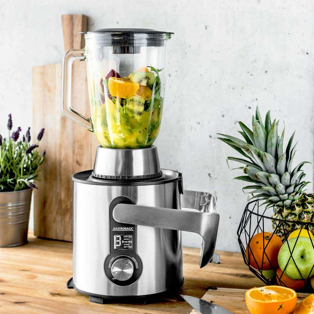 Ein Mixer mit Früchten, auf einer Holztheke. Ananas und Obstkorb im Hintergrund, plus ein Schneidebrett.