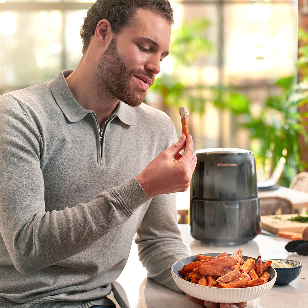 Uomo che tiene un pretzel con salsa, vicino a una ciotola di patatine fritte di patate dolci e una friggitrice ad aria su un tavolo.