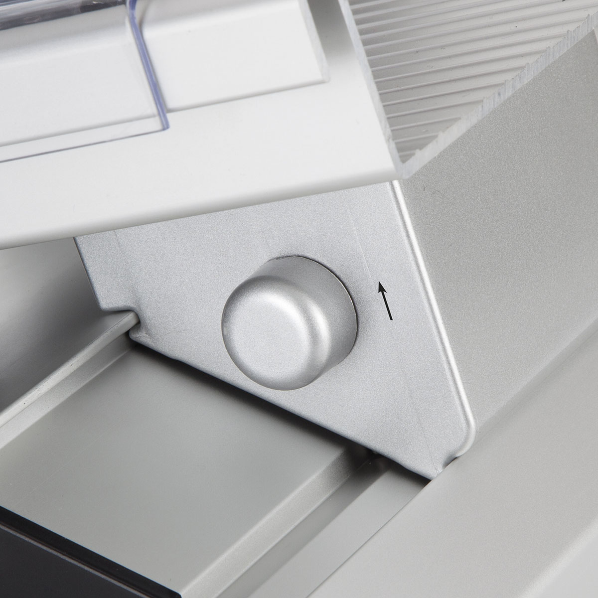 Zbliżenie srebrnego pokrętła na maszynie. Strzałka wskazuje kierunek. Szare i białe powierzchnie w otoczeniu.