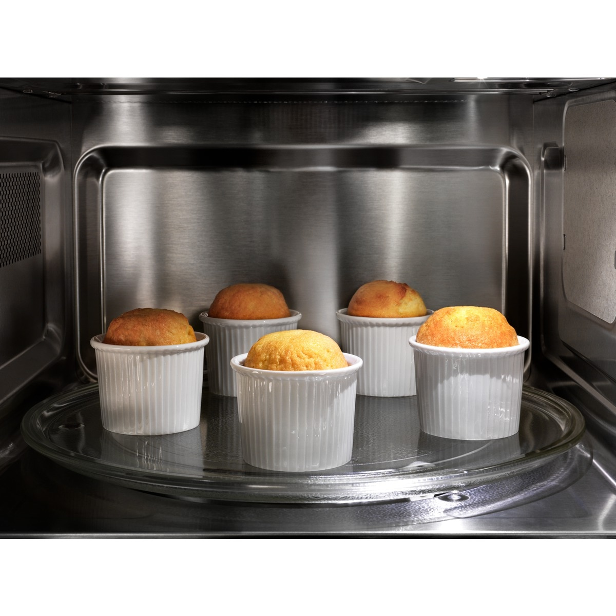 Wewnątrz kuchenki mikrofalowej pięć białych ramekinów z upieczonymi babeczkami na szklanym talerzu.